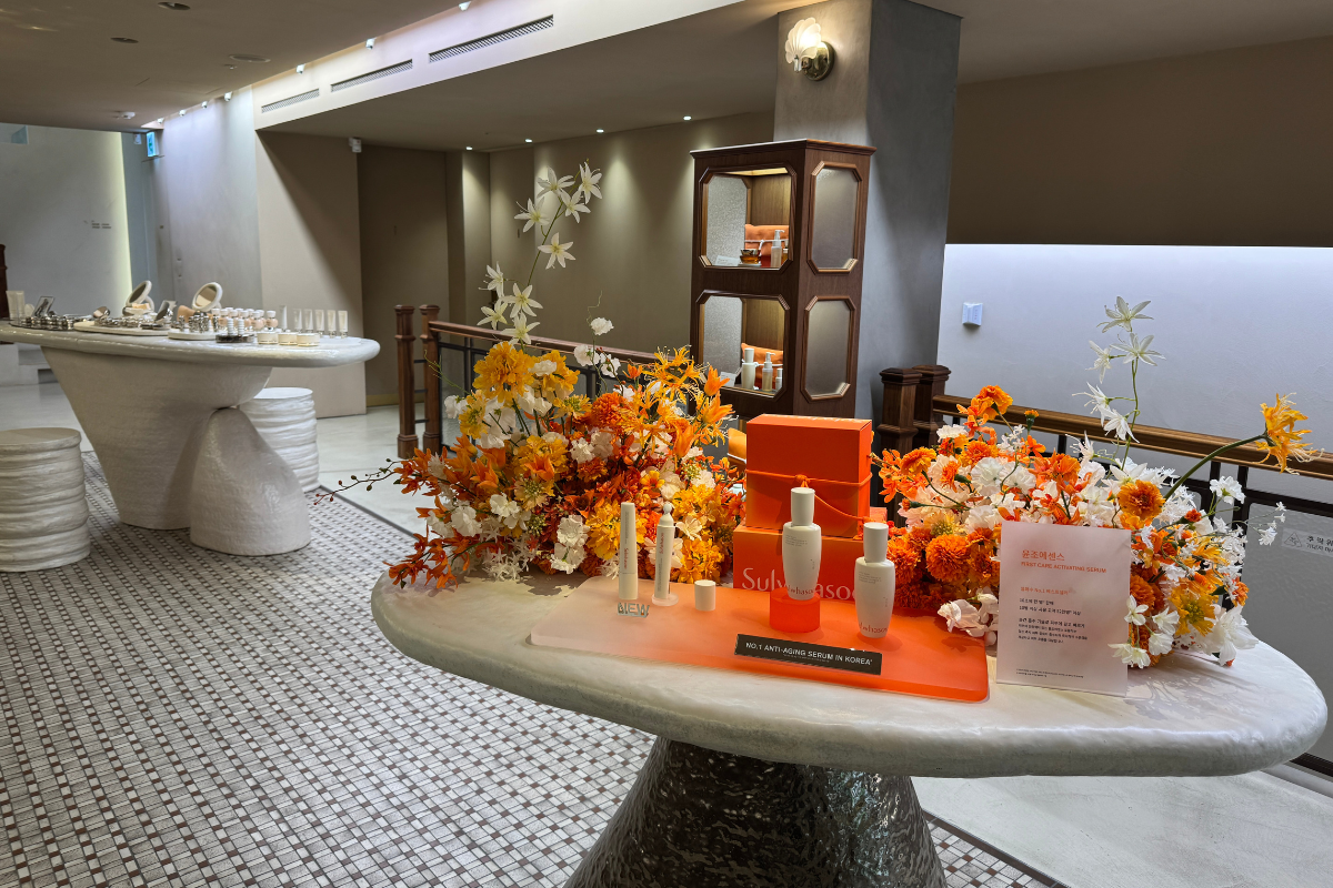 Sulwhasoo Bukchon House nội thất trưng bày với hoa màu cam và trắng, hộp quà thương hiệu, và các chai chăm sóc da trên bàn đá cẩm thạch, với tủ gỗ, ánh sáng dịu, cầu thang, và một khu trưng bày khác ở phía sau.