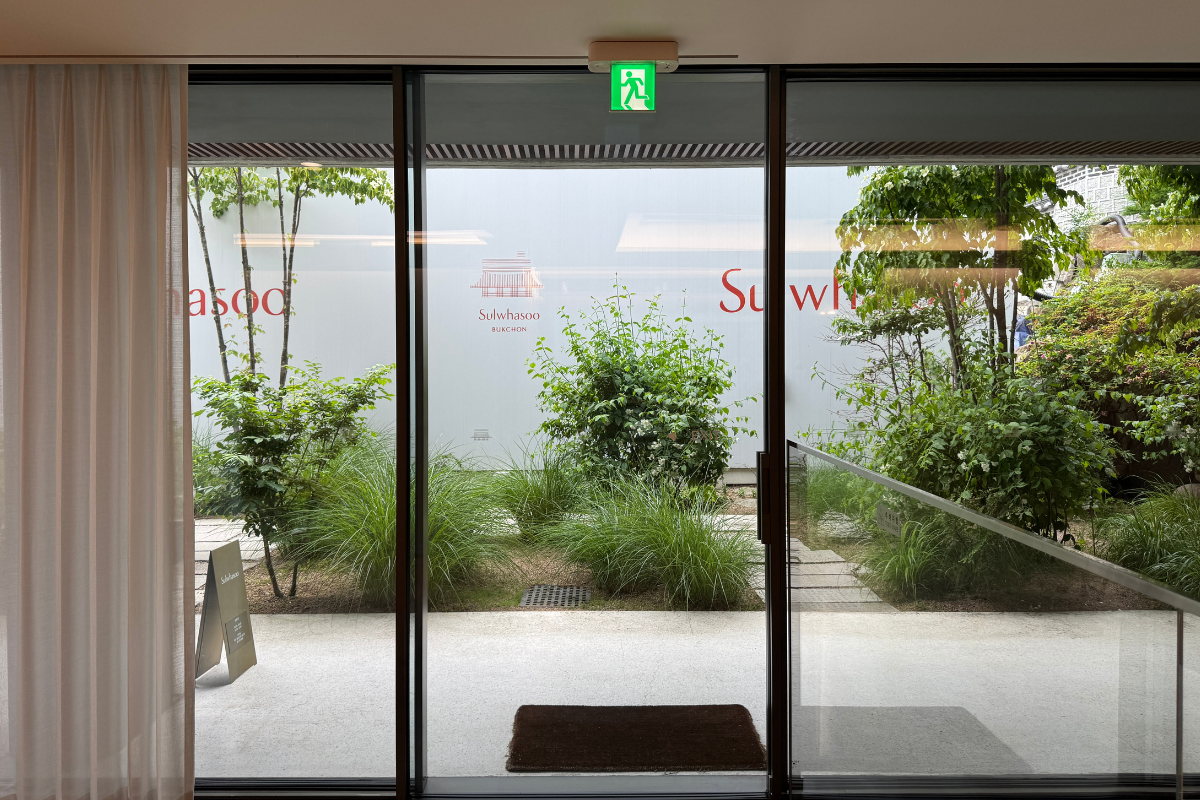 View from inside Sulwhasoo Bukchon House through large glass doors, showing a stone path, small garden, greenery, and the Sulwhasoo logo on the exterior wall.