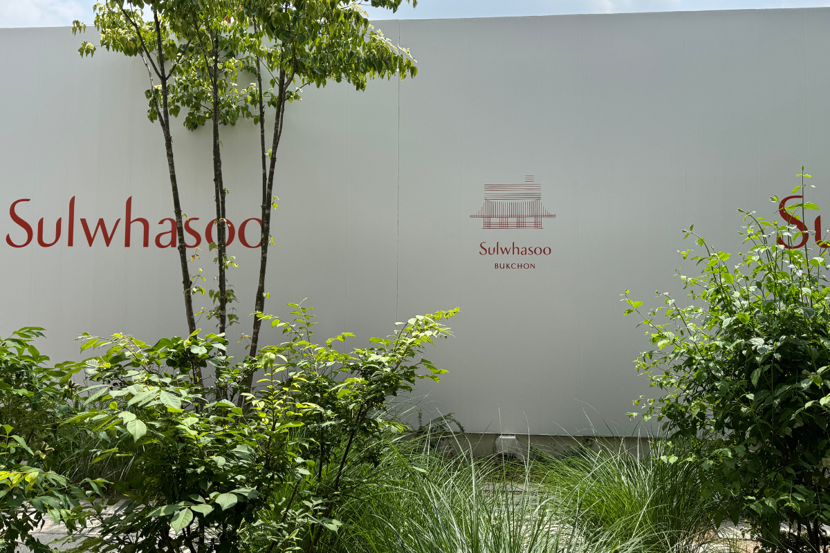 Sulwhasoo Bukchon House sign with red lettering and a hanok icon, surrounded by green garden plants and slender tree trunks.