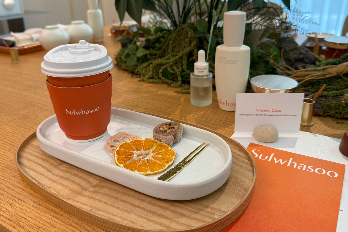 Bố cục quầy tại Sulwhasoo Bukchon House: một cốc thương hiệu màu cam và các sản phẩm chăm sóc da xếp trên khay gỗ, cùng một đĩa sứ trắng đựng lát cam khô, lát củ sen sấy, và các món ăn nhẹ nhỏ, kèm thẻ thông tin thương hiệu, thể hiện trải nghiệm chào đón mang thẩm mỹ thảo dược Hàn Quốc.