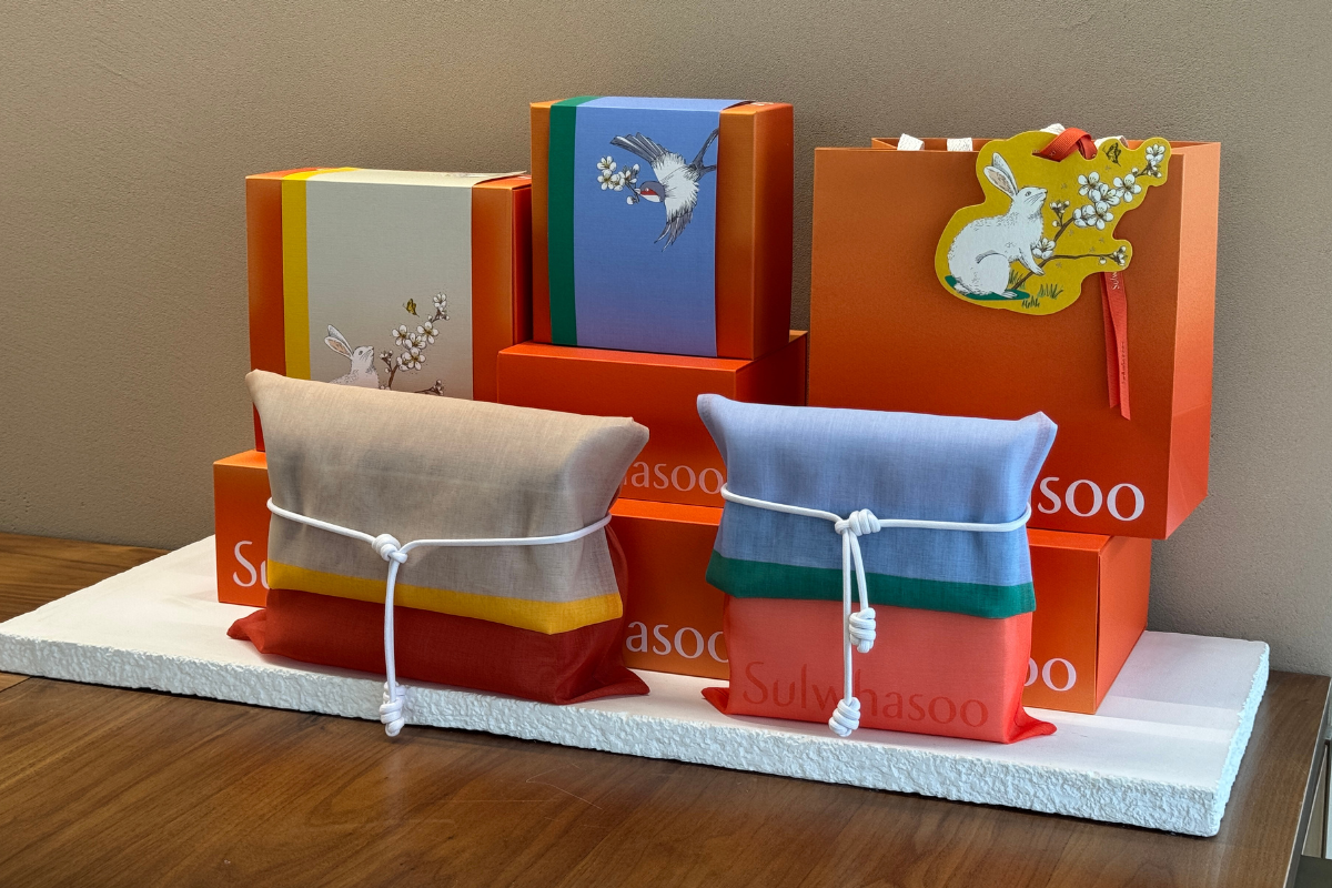 Sulwhasoo Bukchon House display with orange gift boxes, traditional bojagi wrapping pouches, and rabbit and bird illustrations in the background.