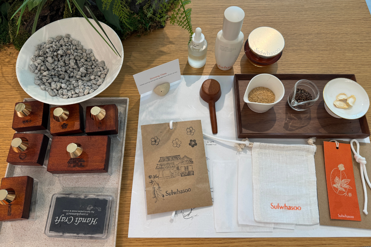 Sulwhasoo Bukchon House DIY table featuring wooden stamps, a stone spoon, ginseng ingredients, skincare bottles, a branded pouch, and orange labels.