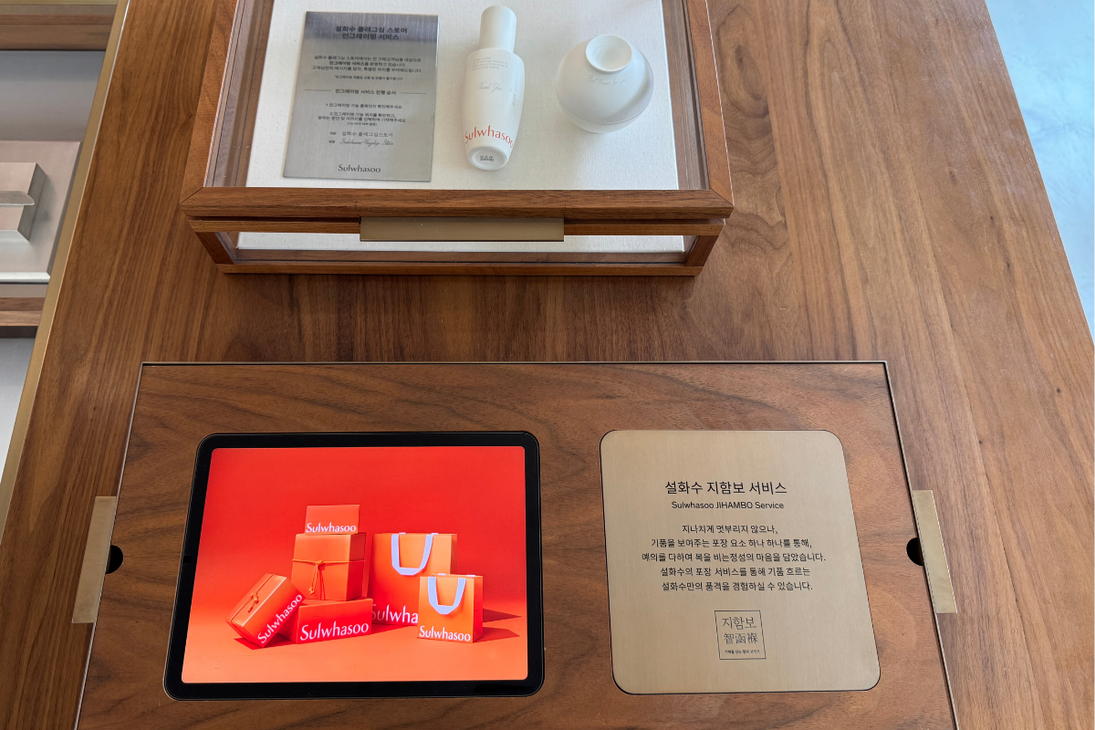Sulwhasoo Bukchon House display table with a tablet showing an orange gift set, skincare bottles, and brand information cards on a wooden surface.
