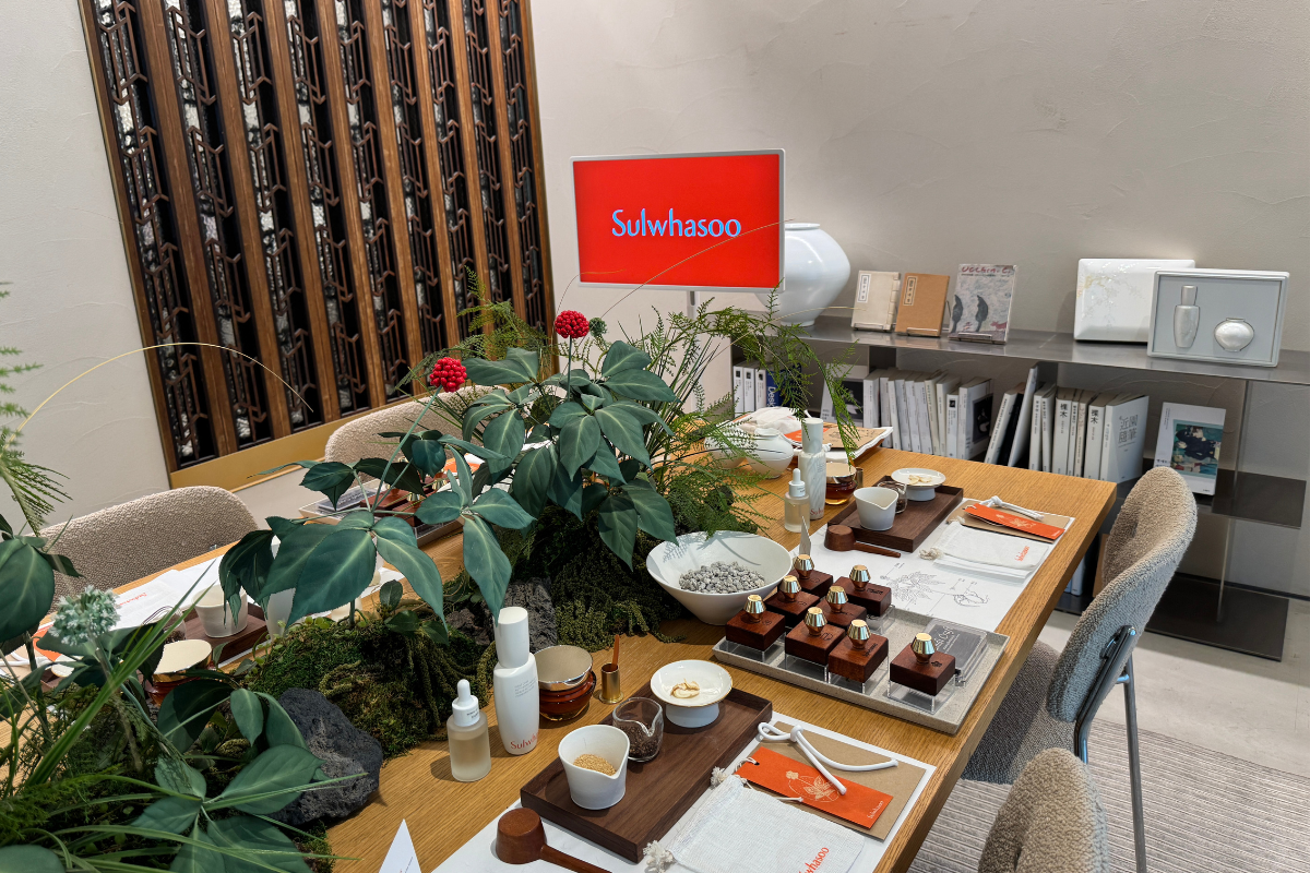 Sulwhasoo Bukchon House experience table with skincare bottles, samples, small porcelain dishes, wooden display stands, packaging, green plants, red berries, a Sulwhasoo logo, and books in the background.