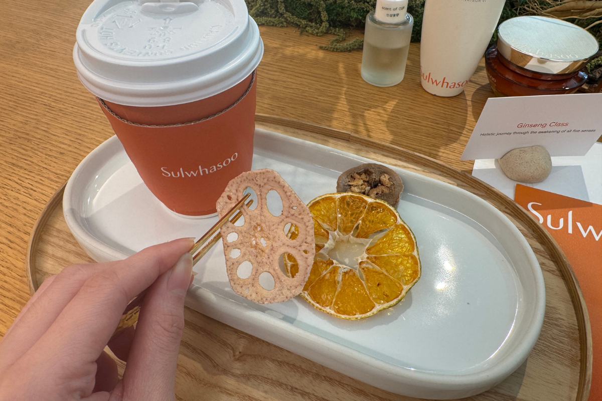 Một cốc thương hiệu màu cam và các mẫu thử Sulwhasoo trên bàn tại Sulwhasoo Bukchon House, cùng đĩa lát củ sen khô, lát cam và các loại hạt, thể hiện bối cảnh tiếp khách thanh lịch kết hợp thẩm mỹ thảo dược Hàn Quốc, trải nghiệm thương hiệu và đồ ăn nhẹ.