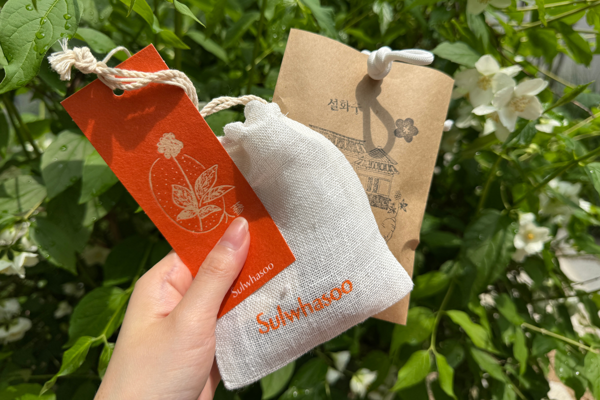 Hand holding a Sulwhasoo drawstring pouch at Sulwhasoo Bukchon House, with an orange branded tag, paper cards, greenery, flowers, and a garden view in the background.