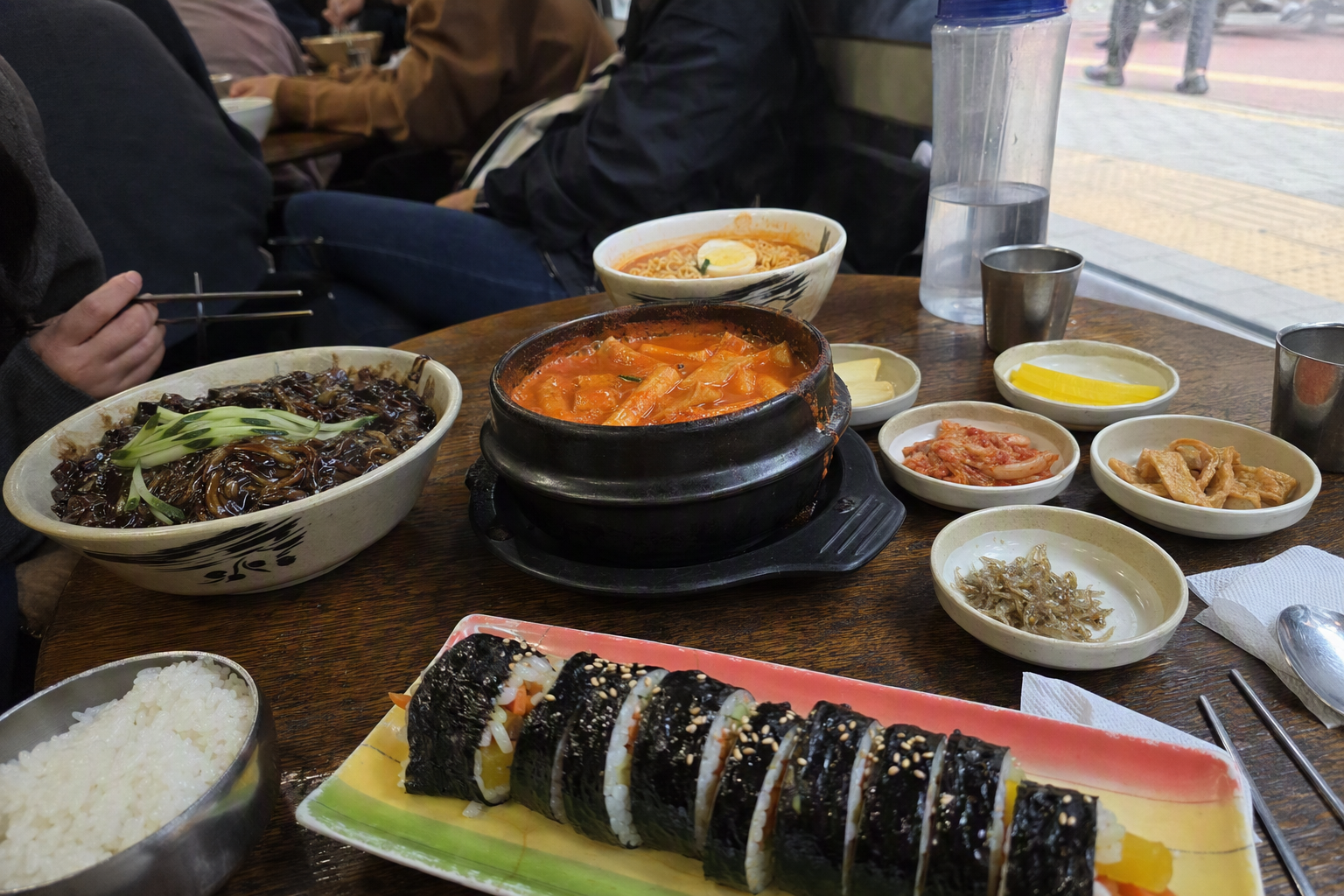 Bright vibrant photorealistic table with kimbap, tteokbokki, jjajangmyeon, ramen, and casual Korean lunch dishes, no Korean text