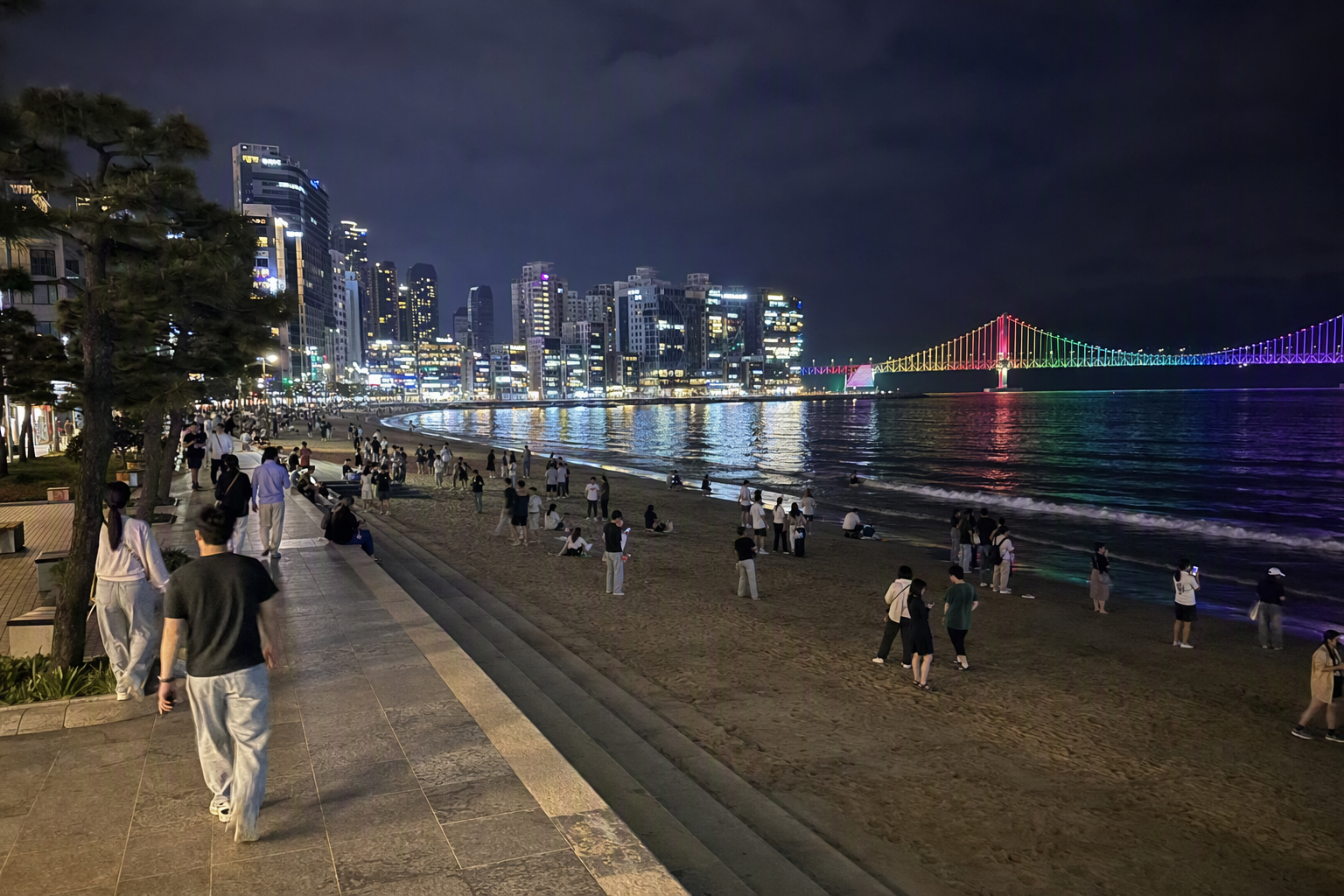 Bright photorealistic night view of Gwangalli Beach with Gwangan Bridge lit in colorful lights, calm water reflections, people walking, no visible text