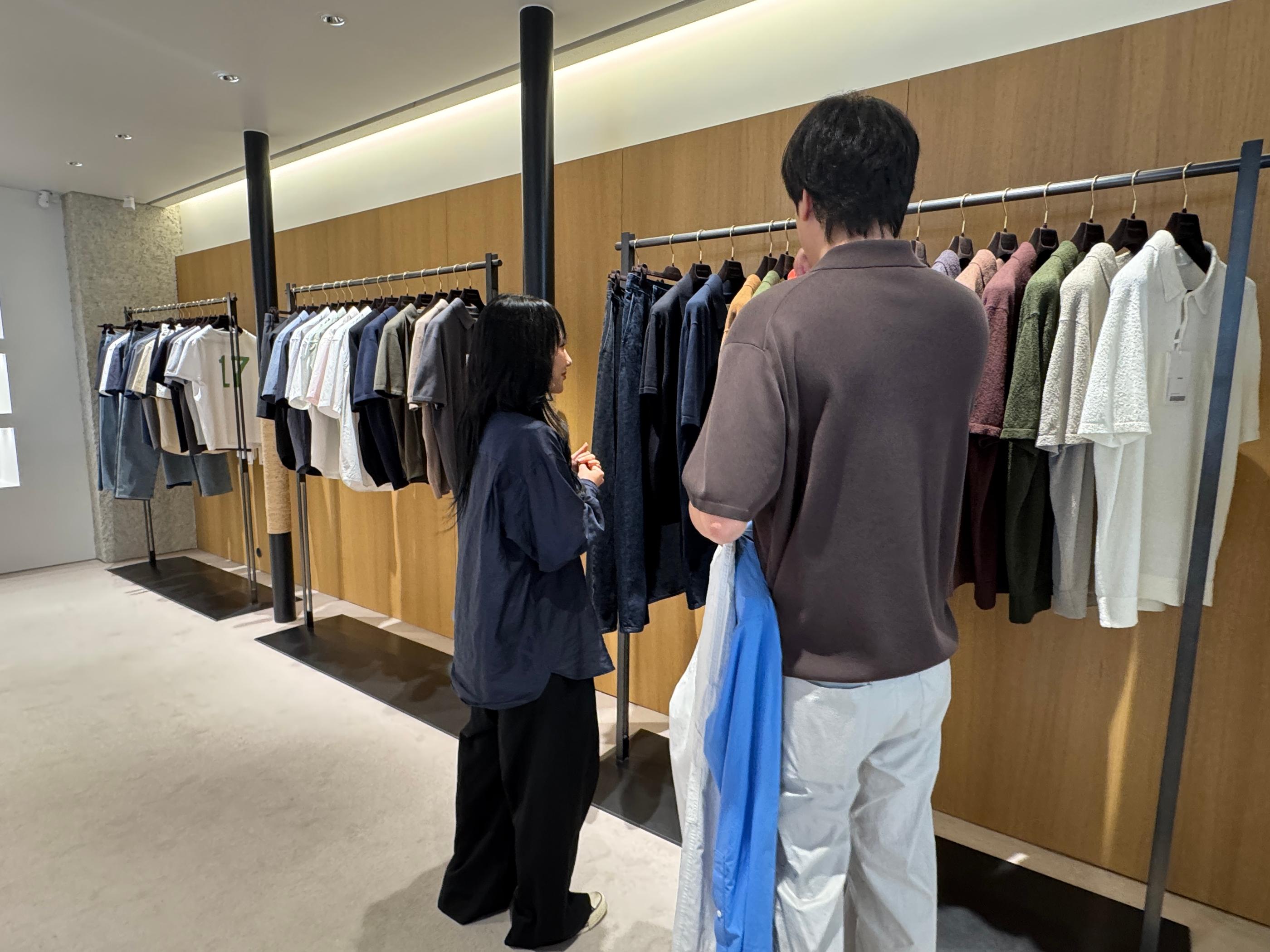 Customers browsing minimalist Pottery menswear racks in the Seongsu store, showing neutral shirts, knitwear and jeans against warm wooden wall panels in a spacious, well-lit interior.