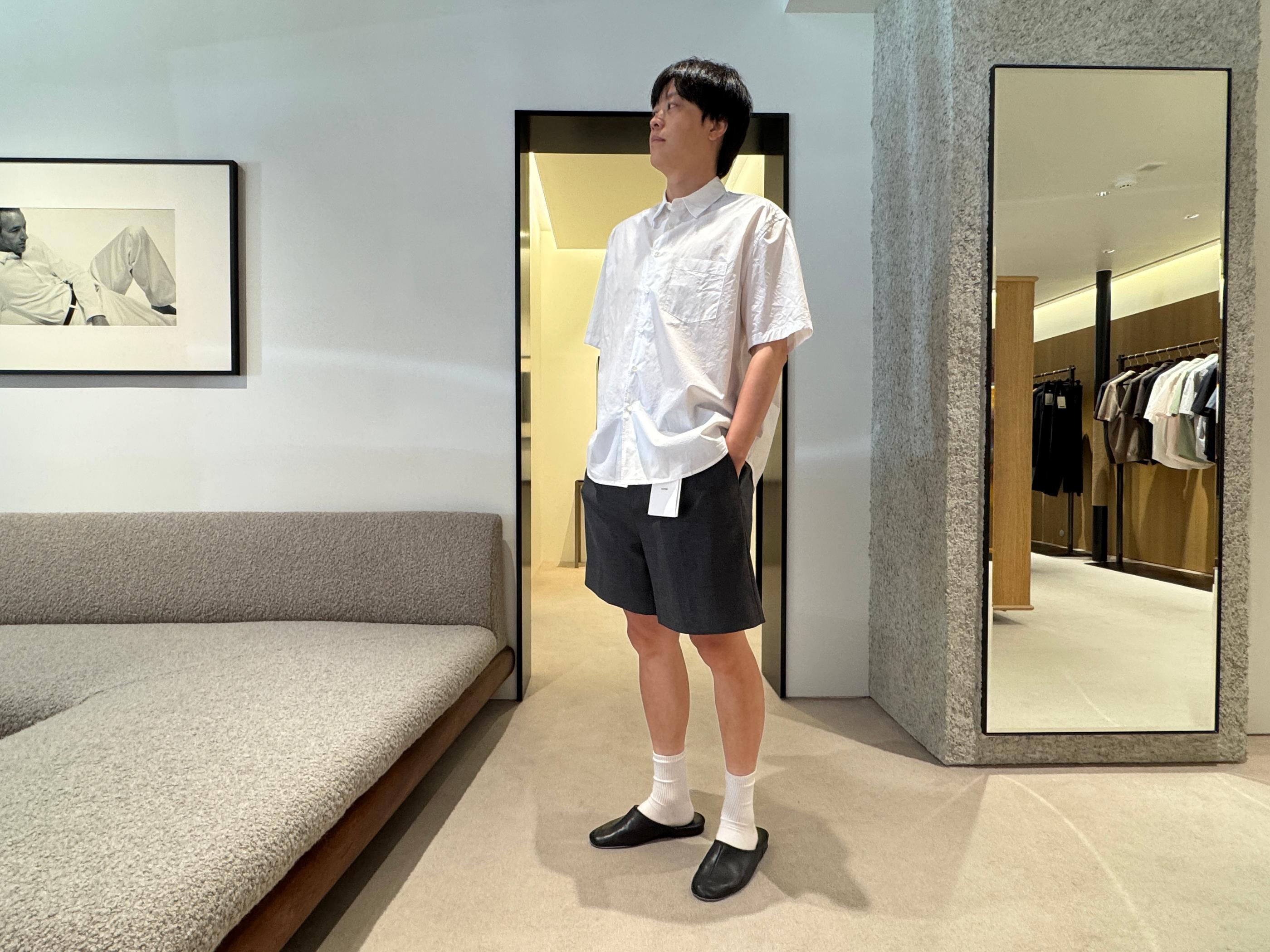 Model in a Pottery Seongsu store wearing a loose white short-sleeve shirt, dark tailored shorts, white socks and black slip-on shoes amid minimalist fittings and a lounge sofa.