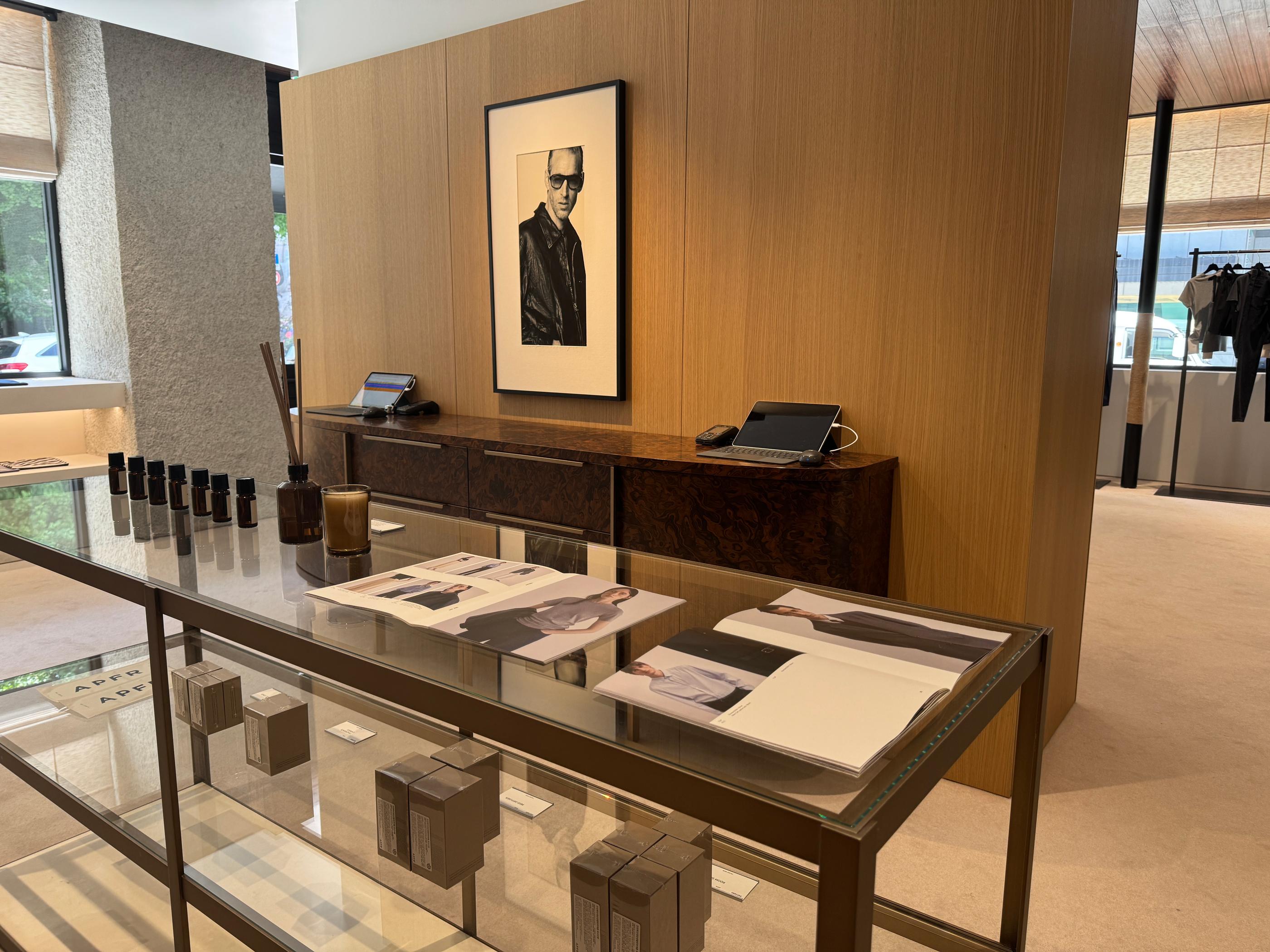 Minimalist Pottery store interior in Seongsu with a glass display table holding catalogs, scent bottles and boxed products, warm wood panels and clothing racks in the background.