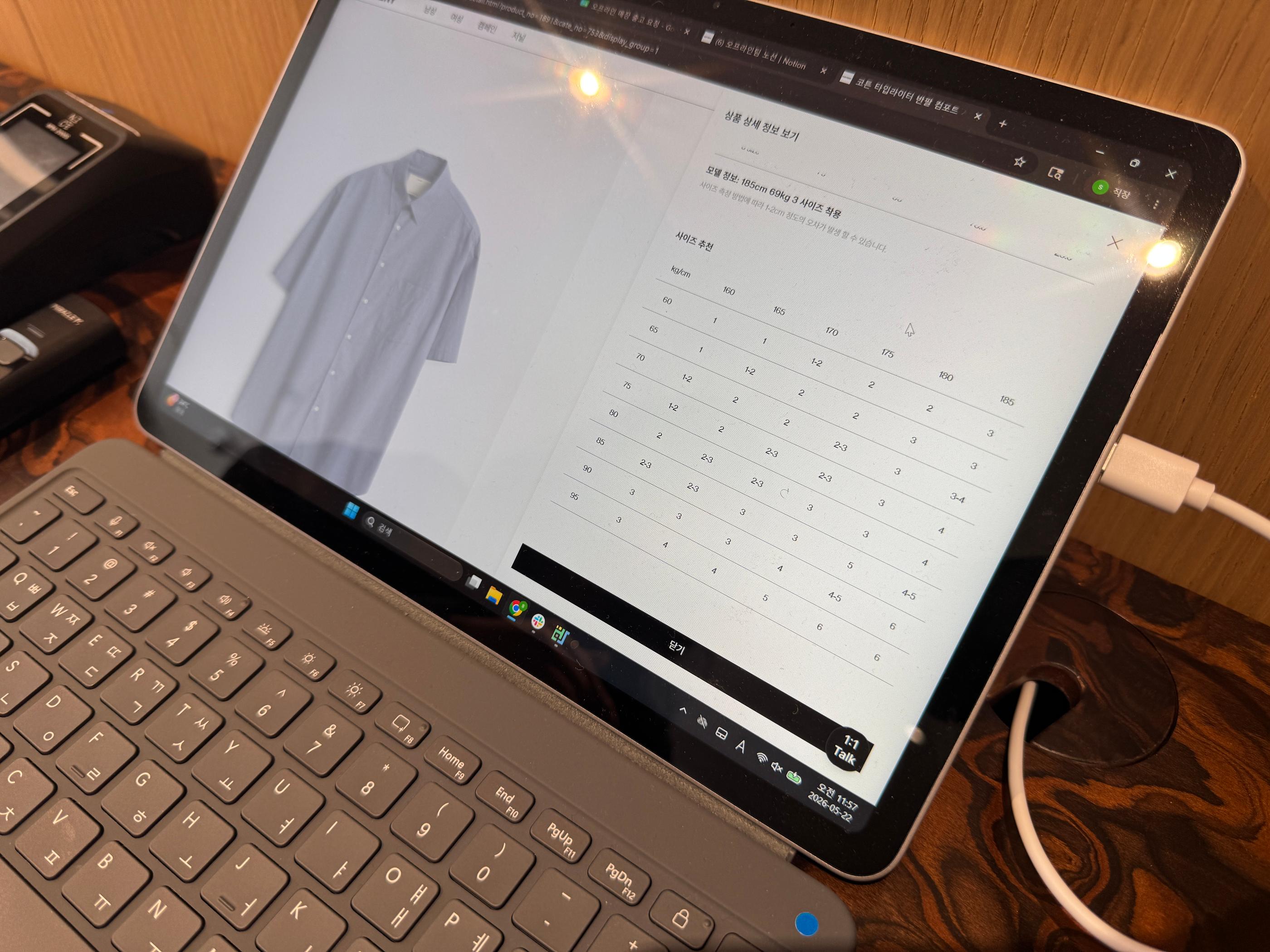 Tablet with keyboard showing Pottery product page and size chart for a short-sleeve shirt, placed on a wooden table with charging cable and card reader—scene from an in-store shopping visit in Seongsu.