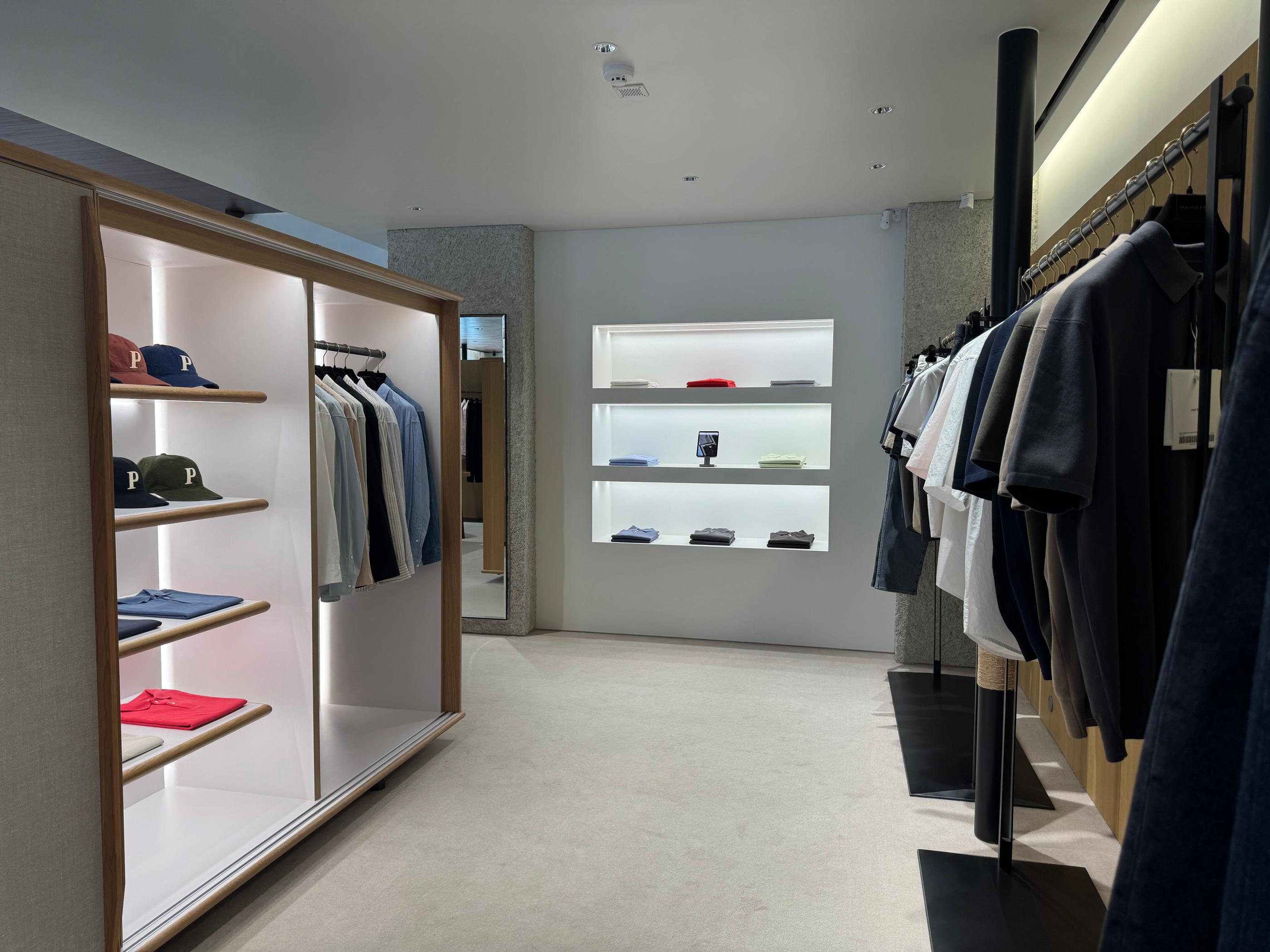 Minimalist Pottery Seongsu store interior with light wood shelving, neatly folded shirts and branded caps on the left, illuminated wall shelves and hanging shirts on the right, calming neutral tones.