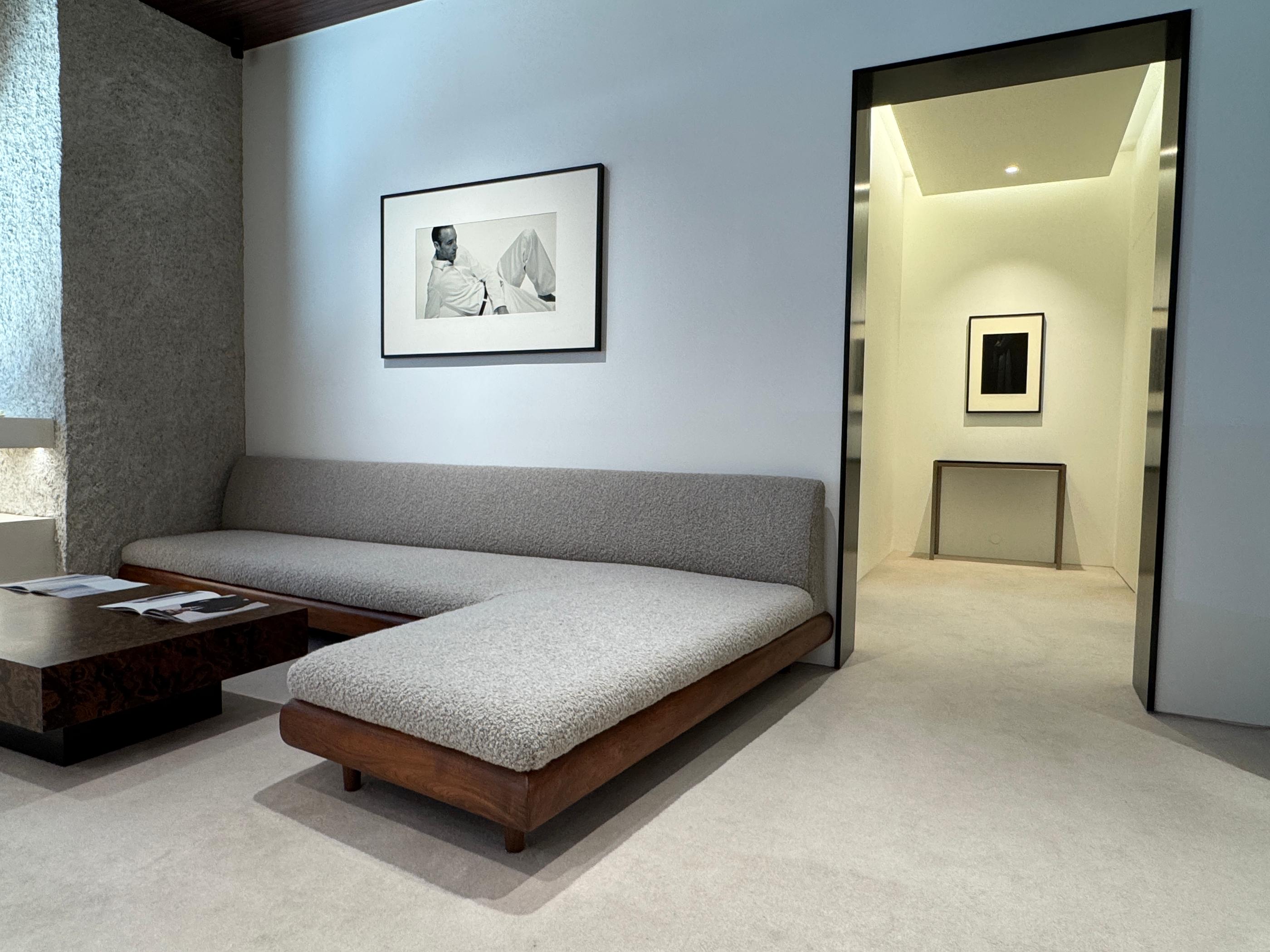 Minimalist Pottery Seongsu store lounge with low wooden framed sofa, textured gray upholstery, wooden coffee table, and a doorway leading to a softly lit fitting area with framed artwork