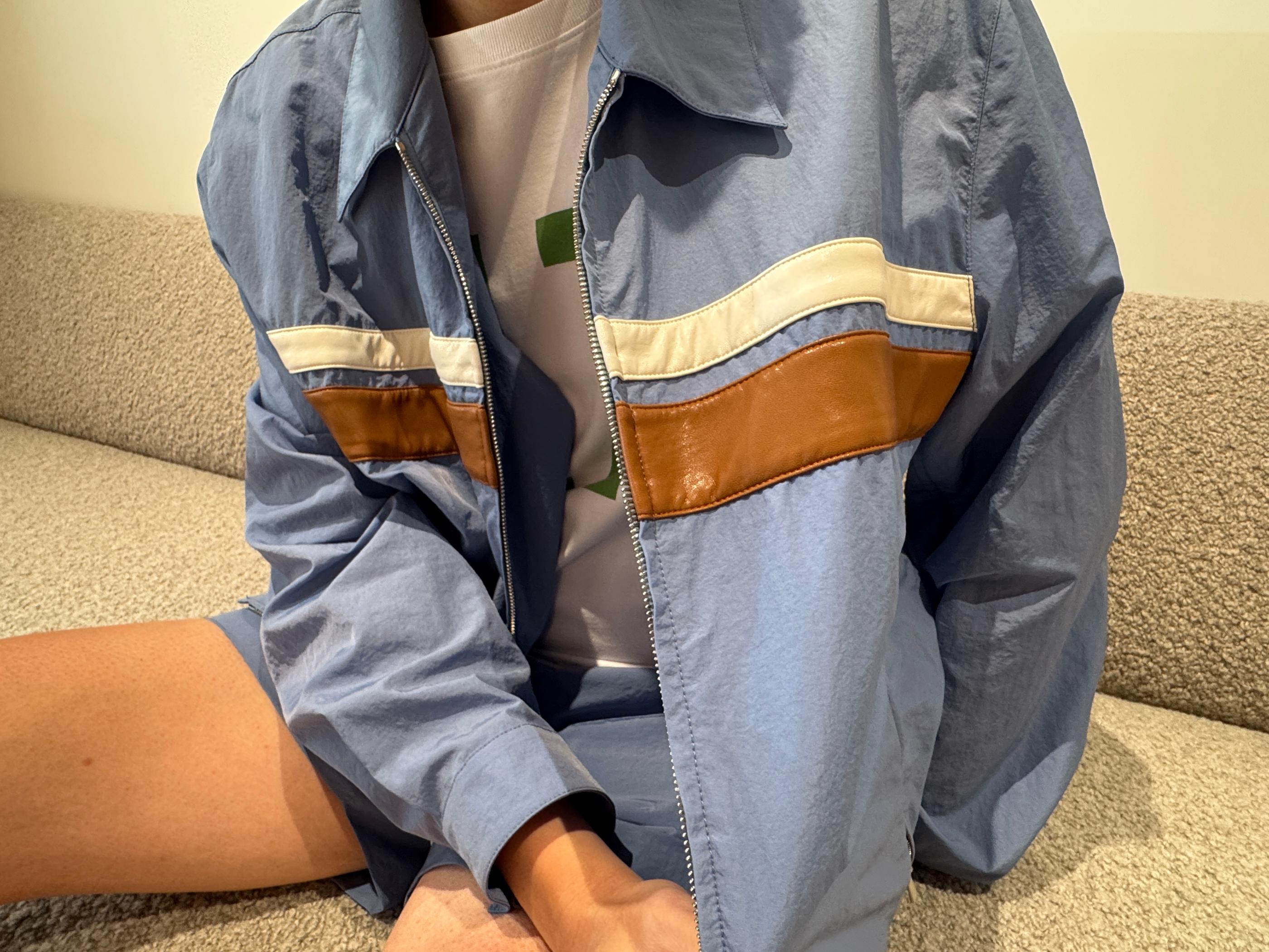 Model seated on a neutral sofa wearing Pottery sky-blue zip jacket and matching shorts with brown and cream color-block stripes, styled over a white graphic tee to show a relaxed Korean menswear summer look.