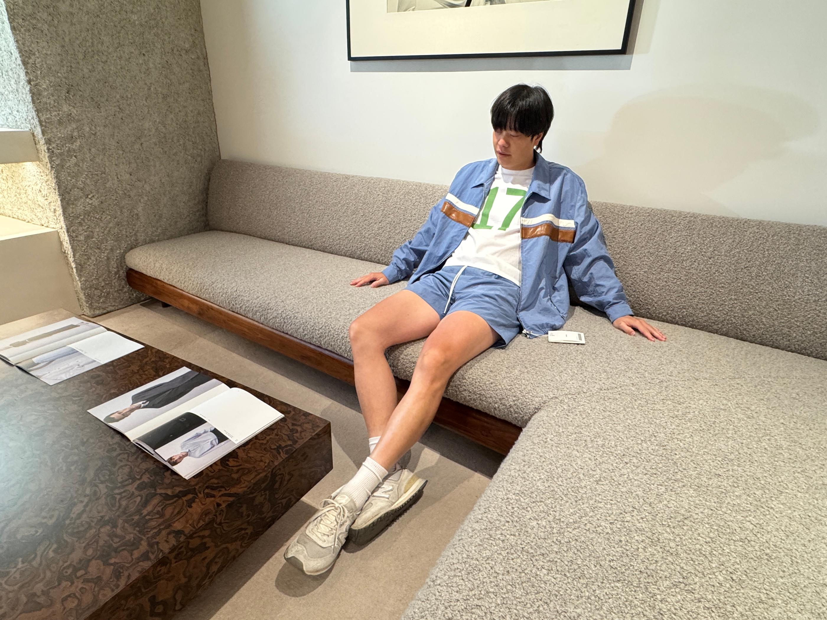 Casual street-style look at Pottery Seongsu: model lounging on a minimalist store sofa wearing sky-blue jacket and shorts set with white graphic tee and sneakers, showing relaxed summer fit.
