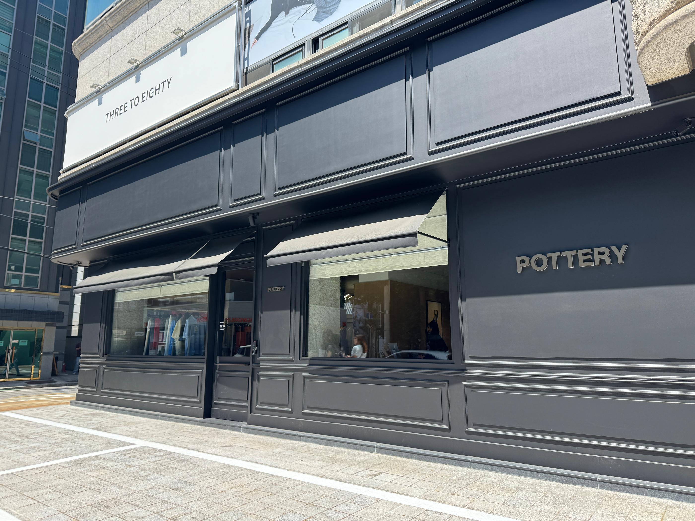 Exterior view of Pottery Seongsu store in Seoul with matte charcoal facade, display windows showing minimalist menswear and the brand name on the wall, sunny street scene.
