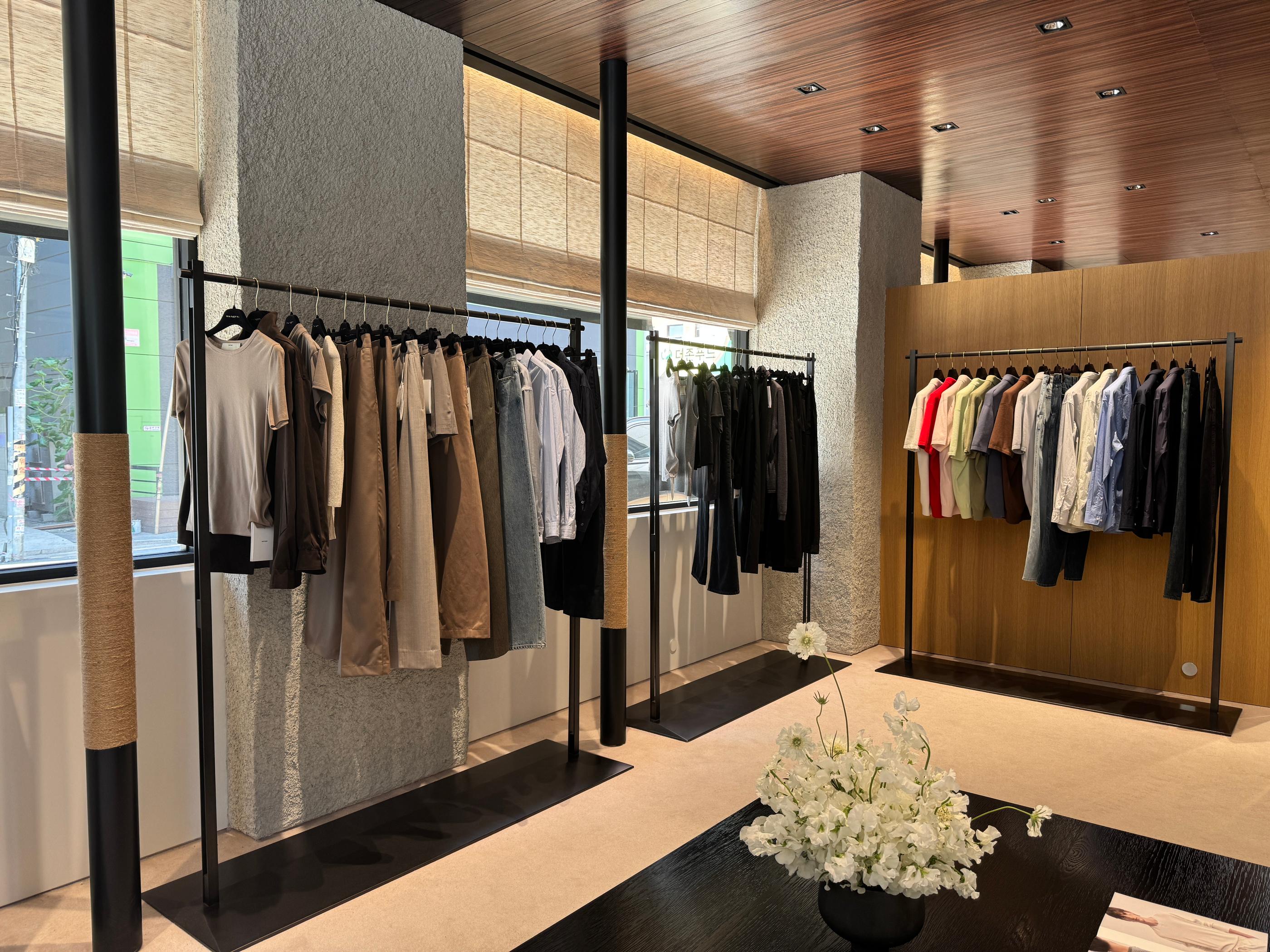 Minimalist Pottery Seongsu store interior with wooden ceiling, clothing racks of neutral and pastel menswear, large windows and a central table with white floral arrangement