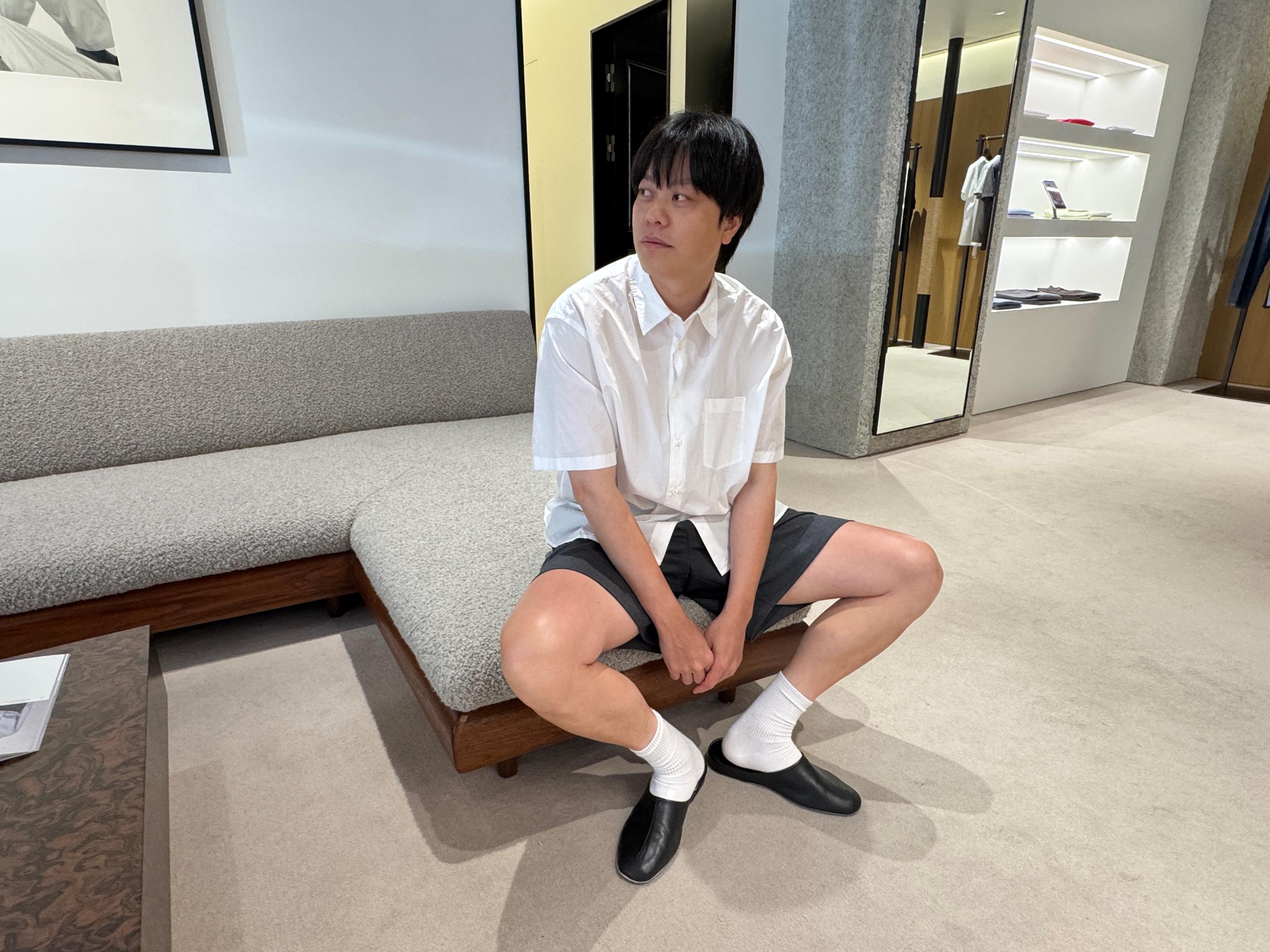 Young shopper wearing a white short-sleeve shirt, gray shorts, white socks and black slip-on shoes sitting on a minimalist wooden sofa inside Pottery Seongsu store, showcasing the brand’s clean, relaxed menswear styling.