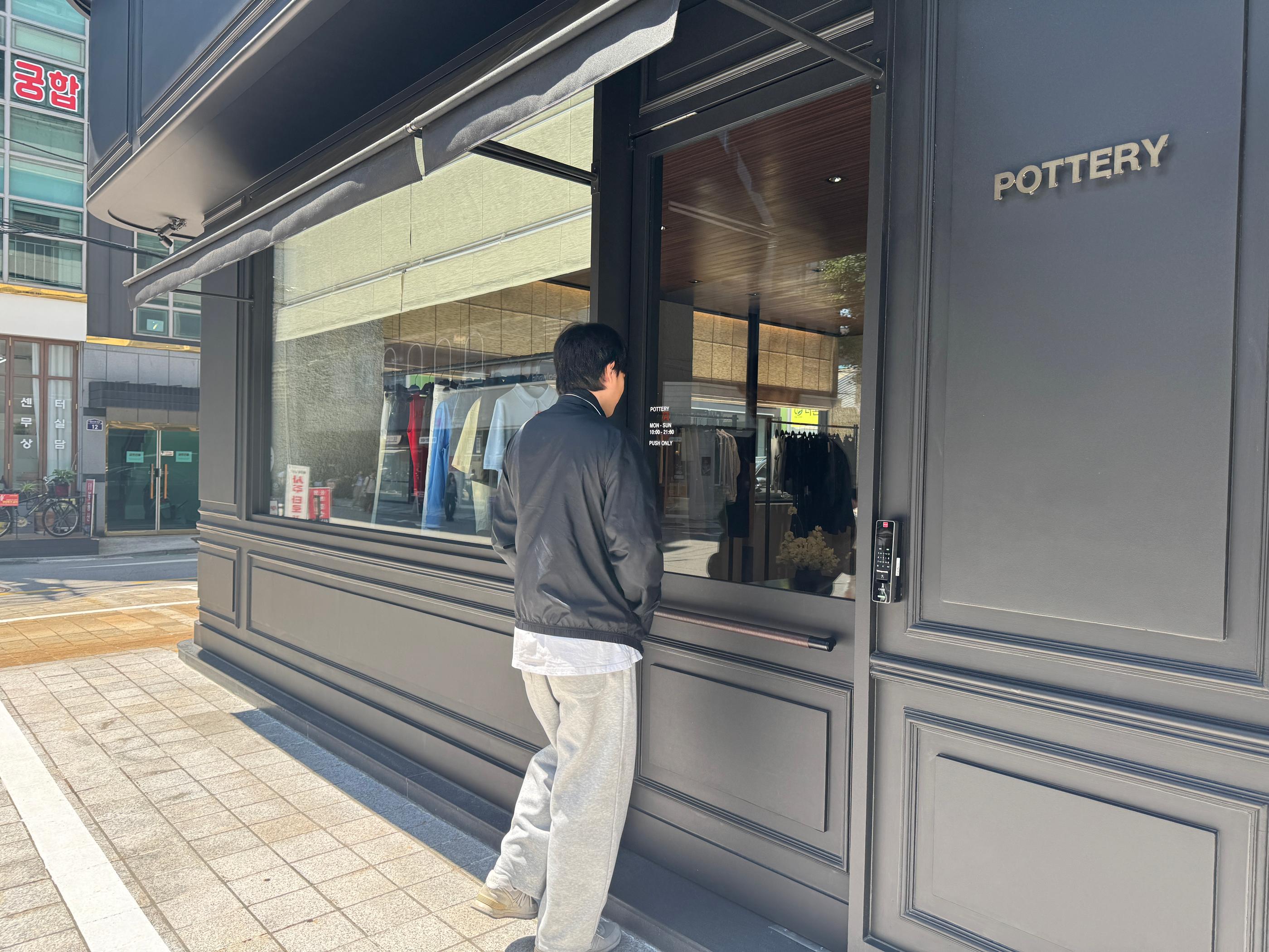 Man peering into Pottery Seongsu storefront on a sunny Seoul street, showcasing the brand’s minimalist dark exterior and clothing displayed in the window.