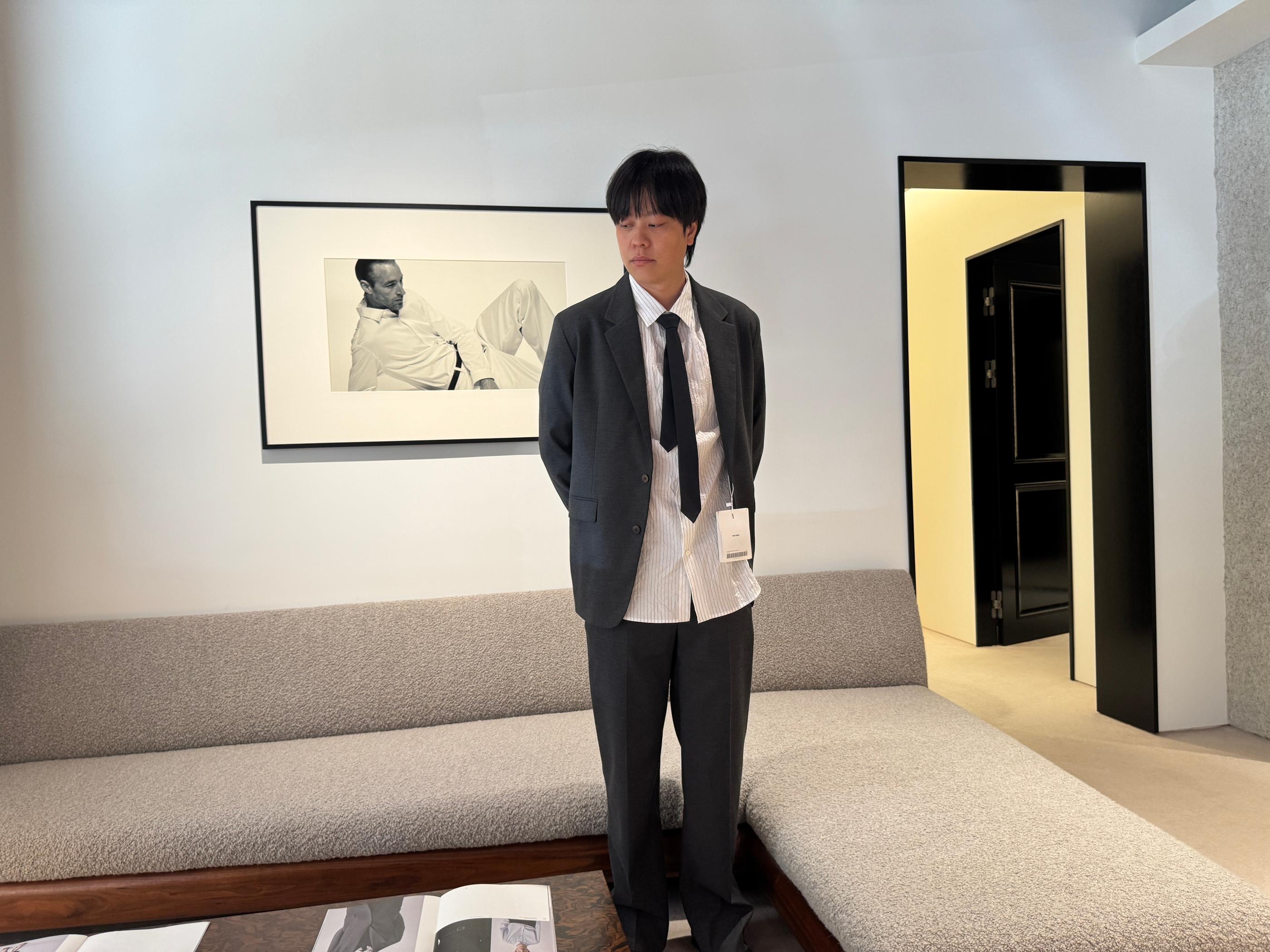 Model trying on a dark gray Pottery suit with striped shirt and loose black tie in the Seongsu store fitting area, minimalist sofa and framed photo visible in the retail space.