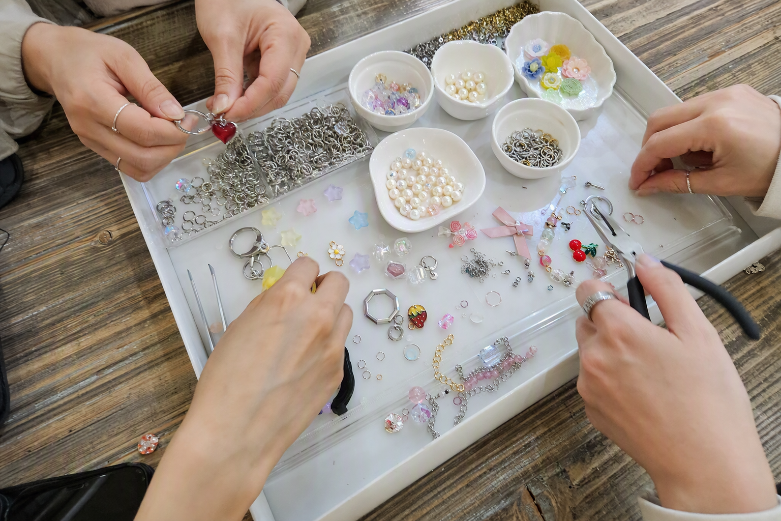 Bright photorealistic overhead view of hands arranging colorful charms, beads, pearls, mini ribbons, and keyring hardware on a white tray, no visible text