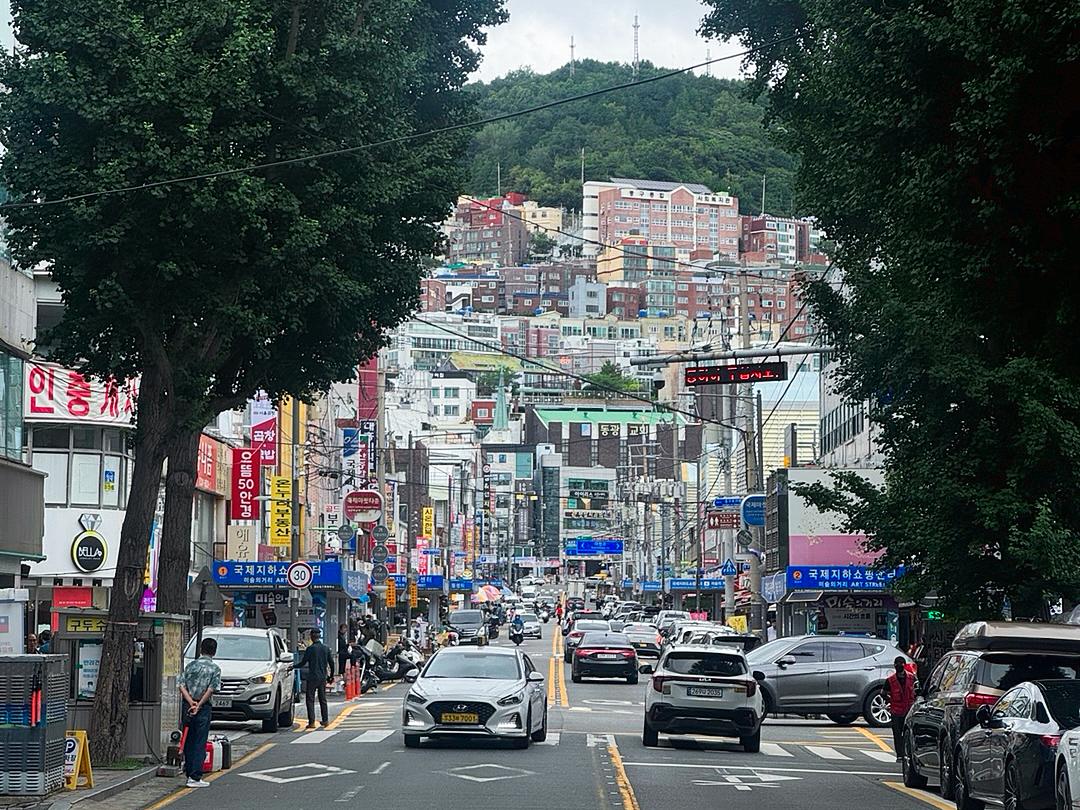 釜山西面街景，透視向上坡的商業街道，人車來往、兩側店家招牌林立，遠處山坡上排列著色彩公寓，顯示西面熱鬧且具城市層次感