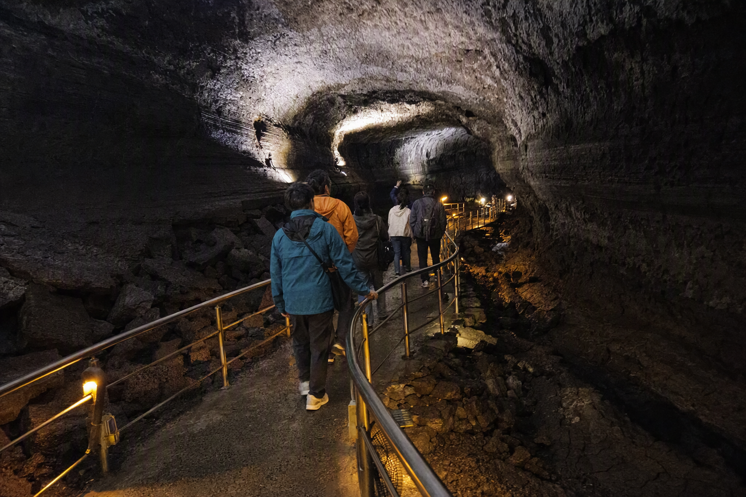 明亮鮮豔、寫實風格的火山熔岩洞內部，岩石紋理強烈，步道照明柔和，遠方有旅客，無可見文字