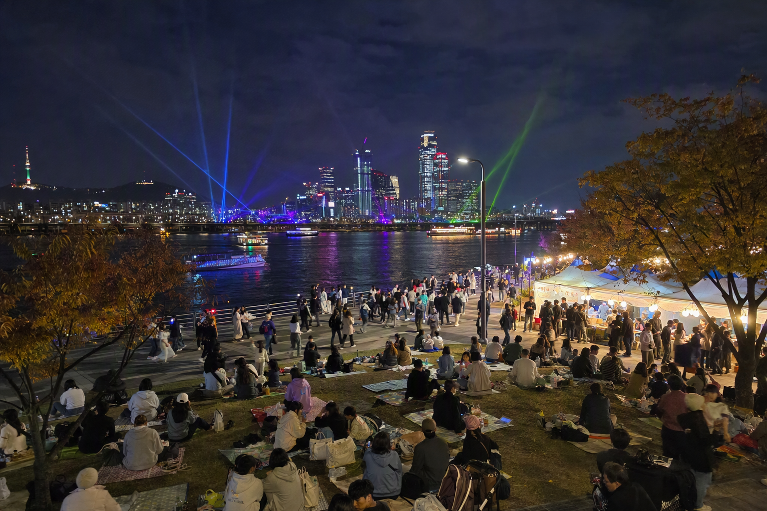 首爾漢江夜間節慶的明亮寫實照片，彩色雷射燈光映照，河岸草地上人潮聚集，城市天際線倒影閃耀，秋夜氛圍熱鬧