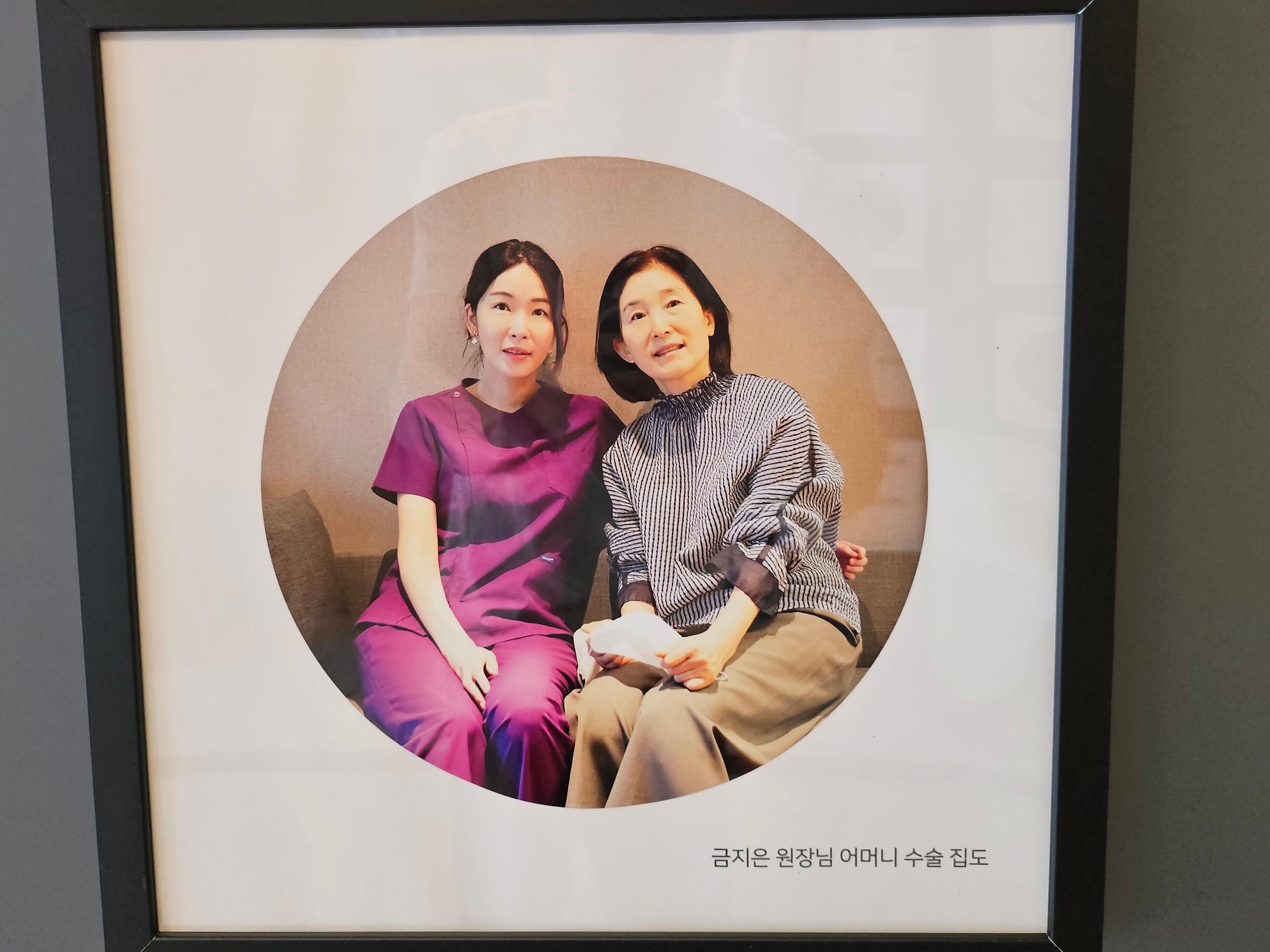 診所牆上相框照片：金智恩院長穿紫色醫療服與媽媽親密並肩坐在沙發上，表情溫和，呈現醫師以家人標準對待患者的故事情感