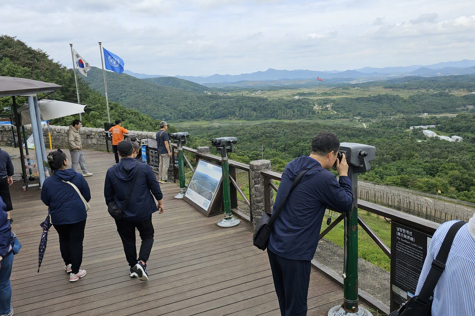 DMZ展望台一帶嘅寫實景觀，遠處有羣山、設有望遠鏡觀景平台、天空平靜，風格莊重如紀錄片式旅遊攝影，無文字