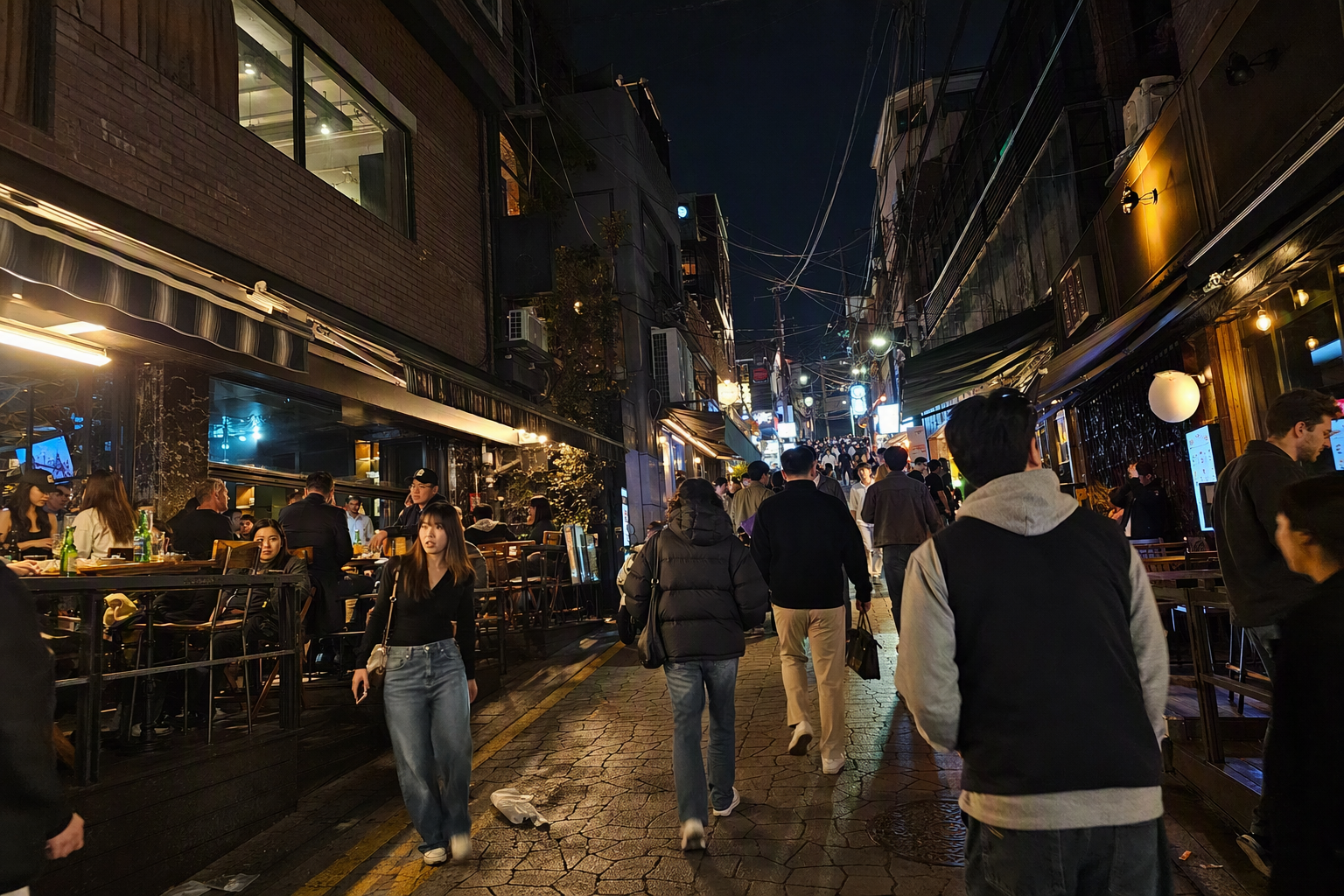 夜晚明亮又充滿活力、寫實風的梨泰院街景，國際餐廳、溫暖燈光、多元行人、斜坡道路，畫面中沒有韓文文字
