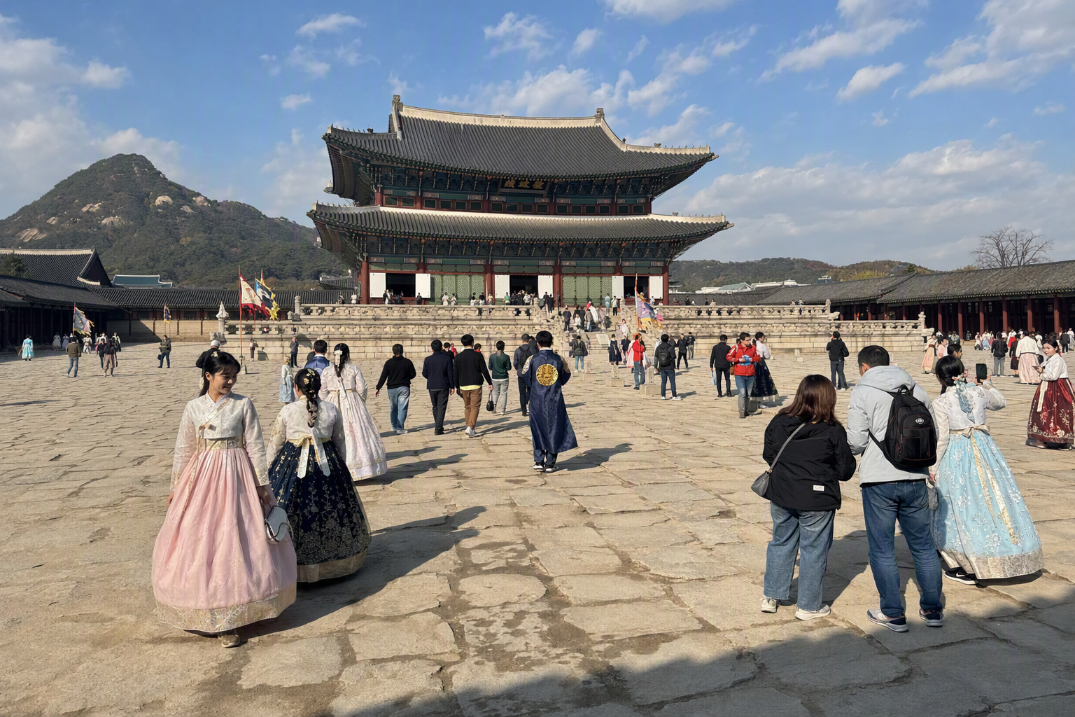 景福宮庭院的明亮鮮豔寫實全景照，有穿著韓服的旅人，背後是群山與藍天，畫面中沒有可見文字