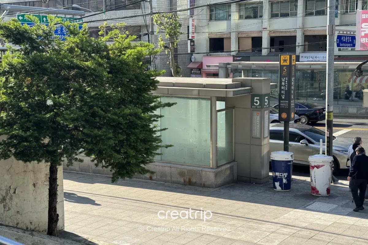 首爾街景:地面出入口標示5號出口、行人和路旁大型飲料廣告桶,旁邊有綠樹與停放車輛,適合說明搭乘長途巴士前確認出口與月台位置的情境