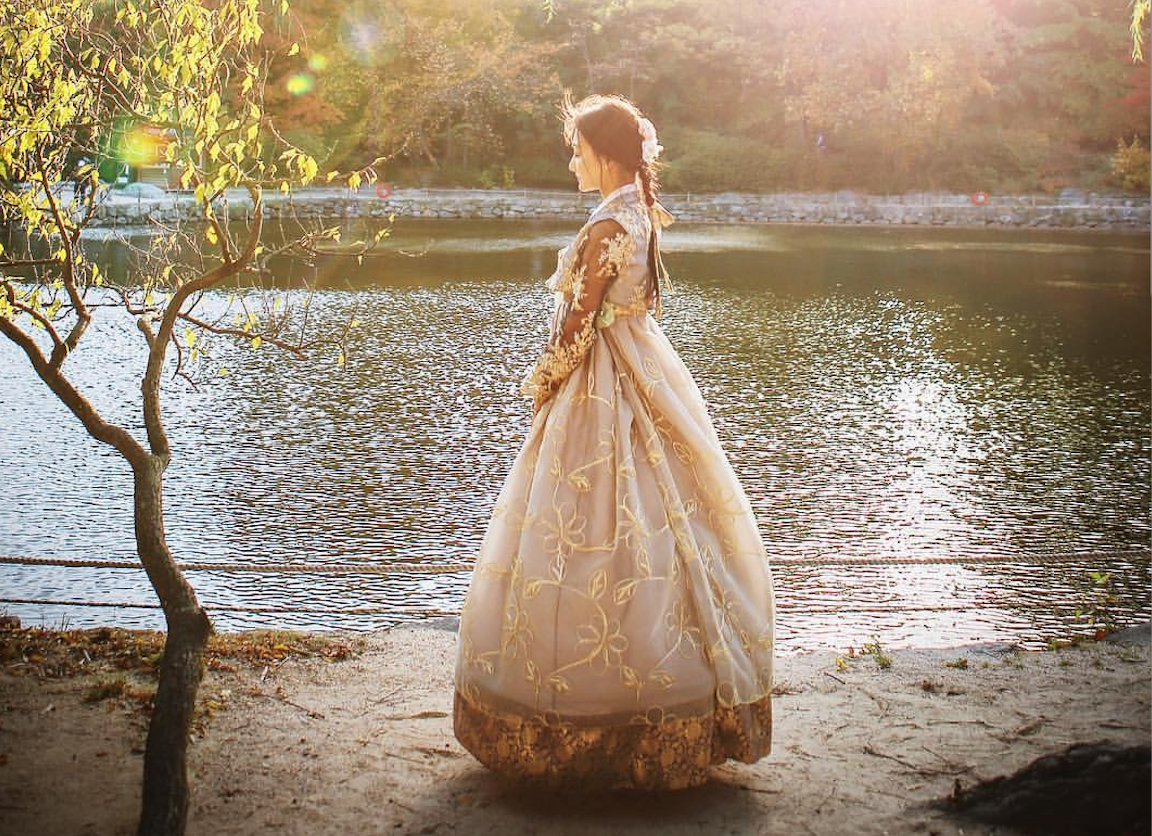 A person wearing a luxurious, ornate hanbok in soft colors with floral embroidery, standing by a serene lake under the warm sunlight.