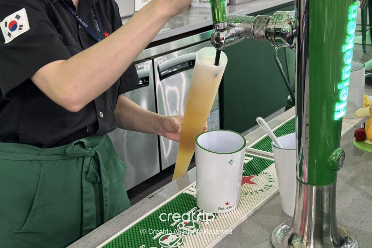 海尼根酒吧吧台场景：服务员在机场酒吧从生啤酒龙头注入大型球棒造型啤酒杯，旁边摆放品牌杯与吧台工具，适合登机前小酌放松的氛围