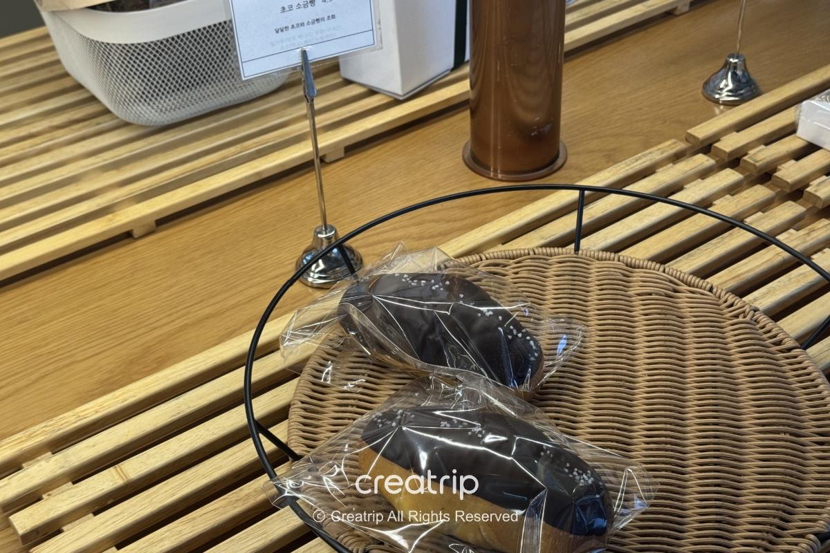 机场面包店展示桌上的两个包装巧克力面包，放在藤编托盘与木质展示架上，标示与CREATRIP浮水印清晰可见，适合抵达或出发时买轻食。