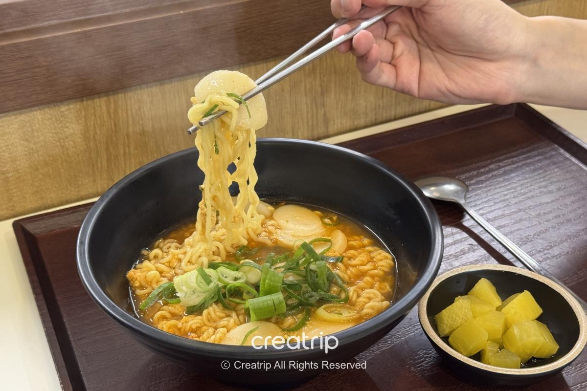 在金海机场餐厅的热腾腾韩式泡面，黑色汤碗内有拉起的面条、葱花与年糕，旁边小碟放著黄色腌萝卜，适合登机前暖胃的一餐。