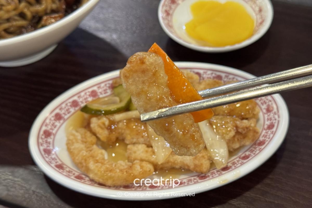 筷子夹起一块糖醋炸猪排与红椒片，下面是小盘糖醋炸物与腌萝卜，背景可见炸酱面碗与木纹餐桌，展现金海机场PLE美食街的韩式中华风味。