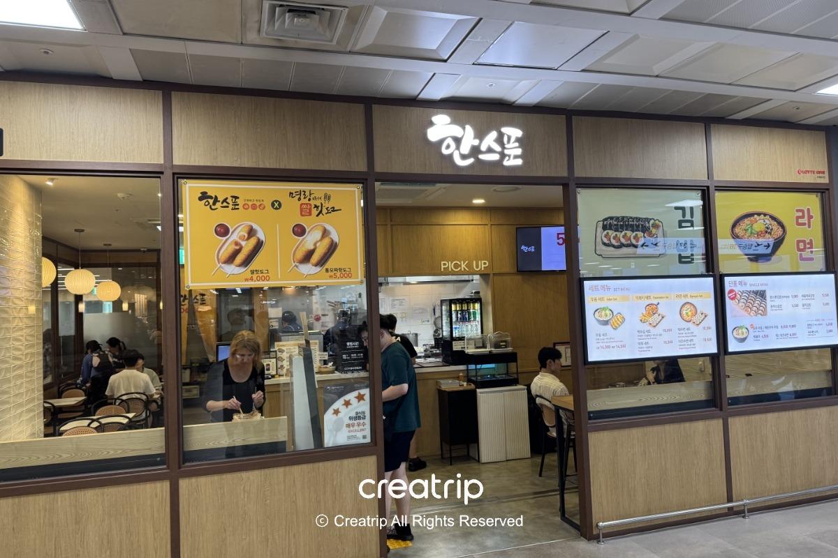 釜山金海機場國內線出發層的HAN SPOON小吃店外觀，木質門面與玻璃櫥窗可見內用座位、點餐與取餐櫃檯，以及菜單照片與客人用餐情形，適合登機前快速享用年糕、飯捲等韓式小吃。