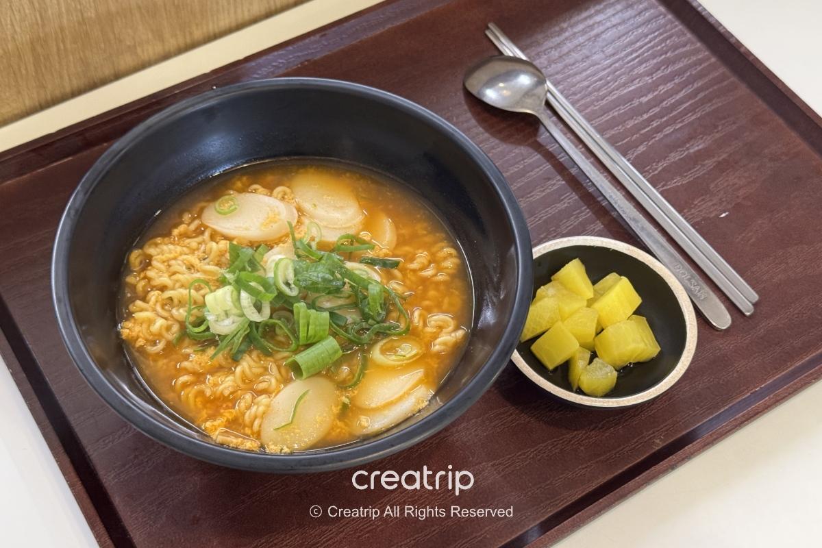 釜山金海机场HAN SPOON的辣炒年糕泡面套餐，黑碗装热汤面配鱼板与葱花，旁边小碟放著黄色腌萝卜，餐盘上有筷子与汤匙，适合登机前快速暖胃。