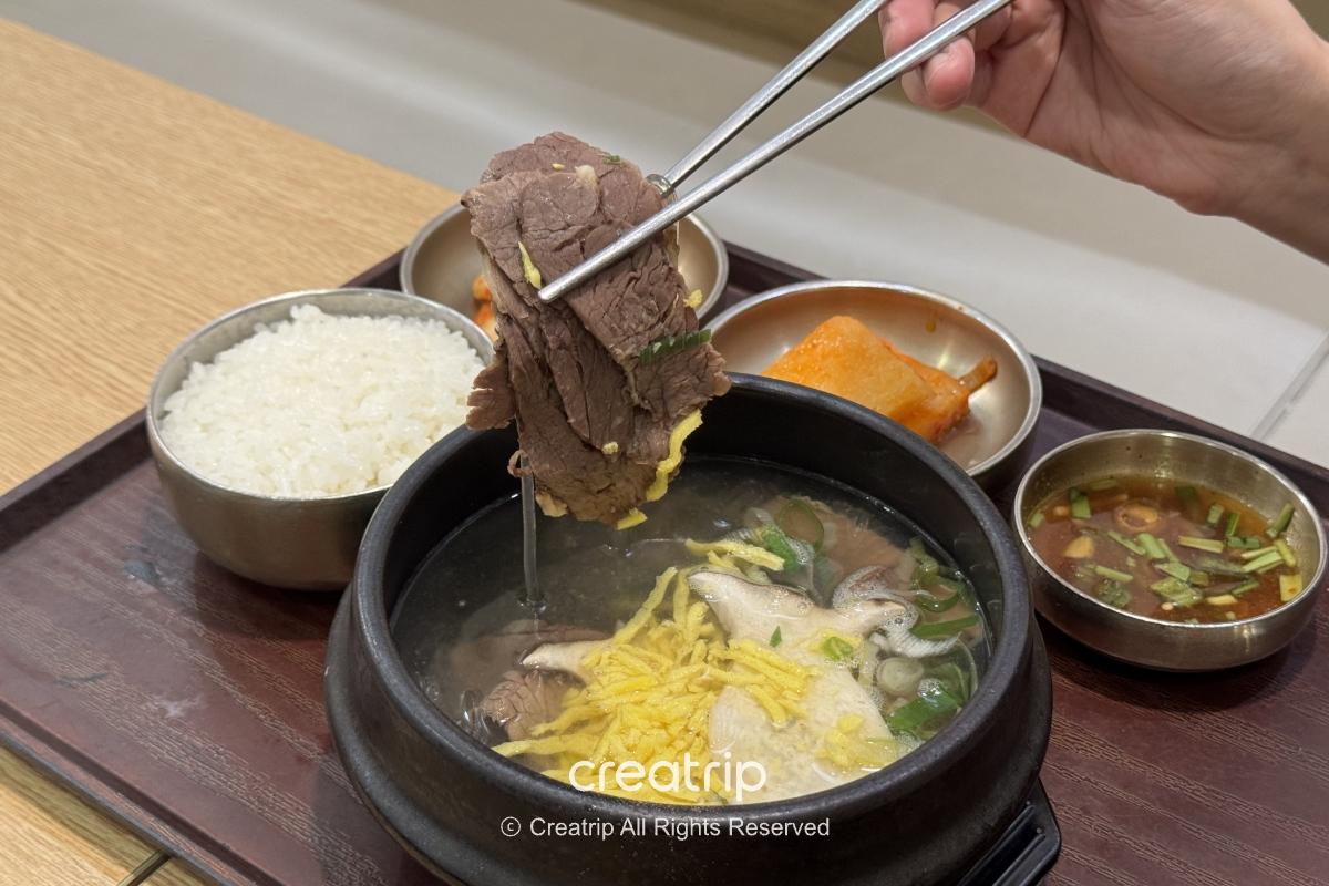 金海机场孝子牛骨汤的牛胸肉牛骨汤套餐画面：黑陶锅热汤搭配厚切牛胸肉、蛋丝与香菇，附白饭、小菜与酱料，正用筷子夹起一片牛肉准备吃。