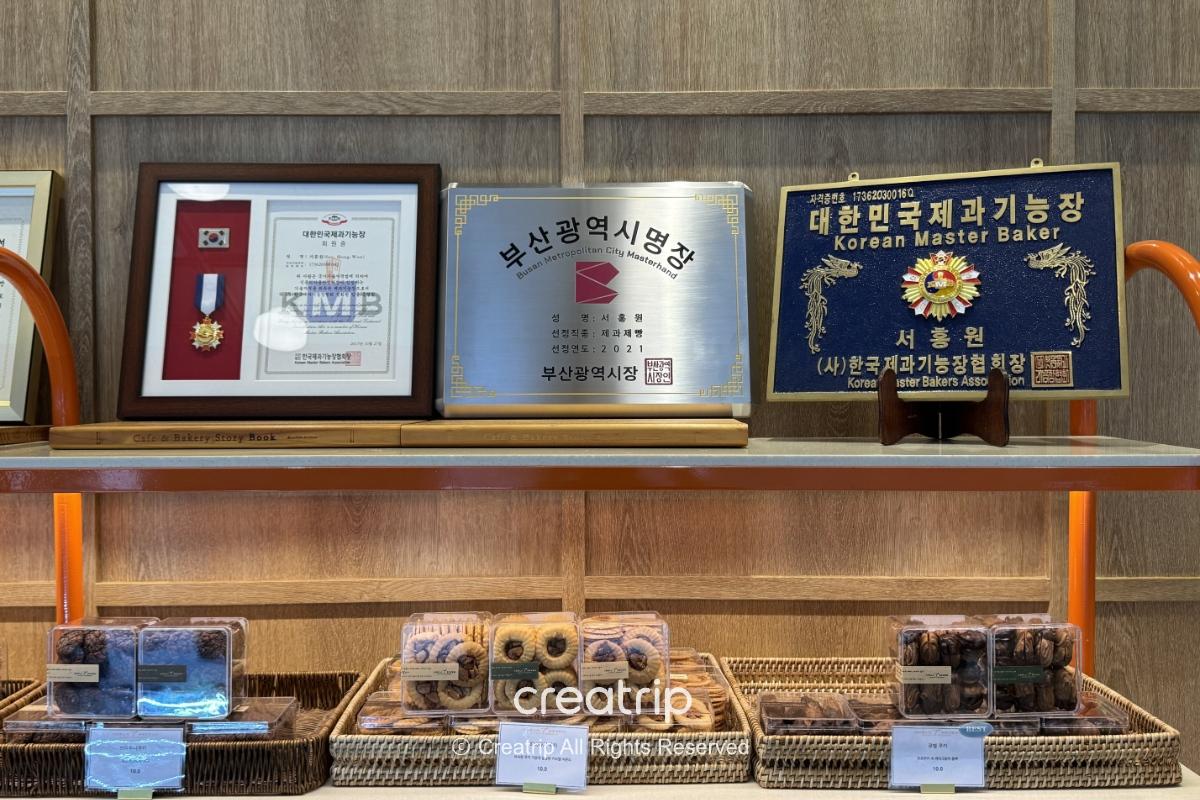 釜山金海机场ADELA 7面包店展示架：上层摆放奖牌与证书，下层有精美包装的饼干与糕点，适合当伴手礼或机场小点心。