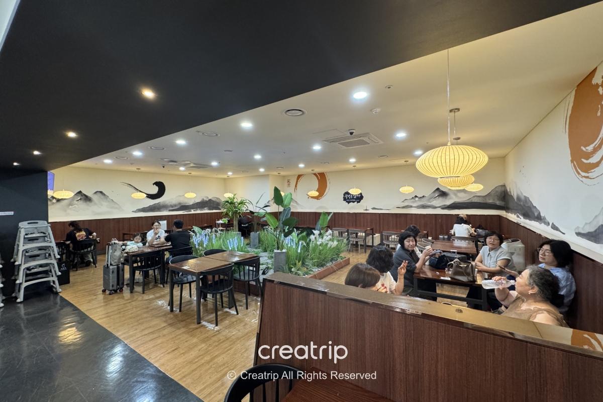 釜山金海机场内的明亮用餐区，木质桌椅与绿植隔间，旅客带著行李或家庭聚餐，墙面水墨山景与温暖吊灯营造舒适候机用餐氛围。