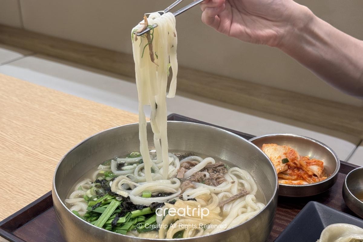 在釜山金海机场孝子牛骨汤的牛肉刀削面套餐特写：金属碗中热腾腾的汤面、大片牛胸肉、海苔与葱花，旁边有小碟泡菜与配料，筷子夹起柔嫩面条准备品尝。
