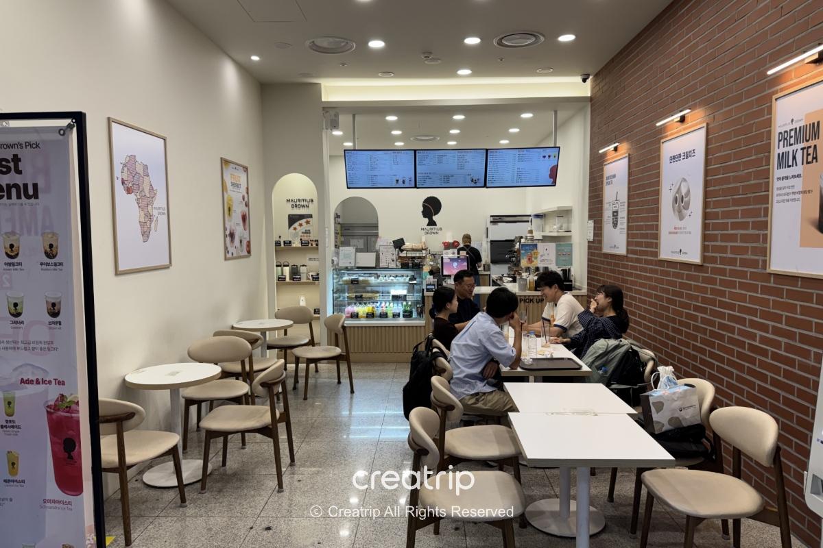 釜山金海机场Mauritius Brown饮料店内景，浅色简约座位与墙面菜单，旅客于桌边聊天、柜台显示器与冷藏柜展示饮品甜点，适合抵达后买杯奶茶或内用休息