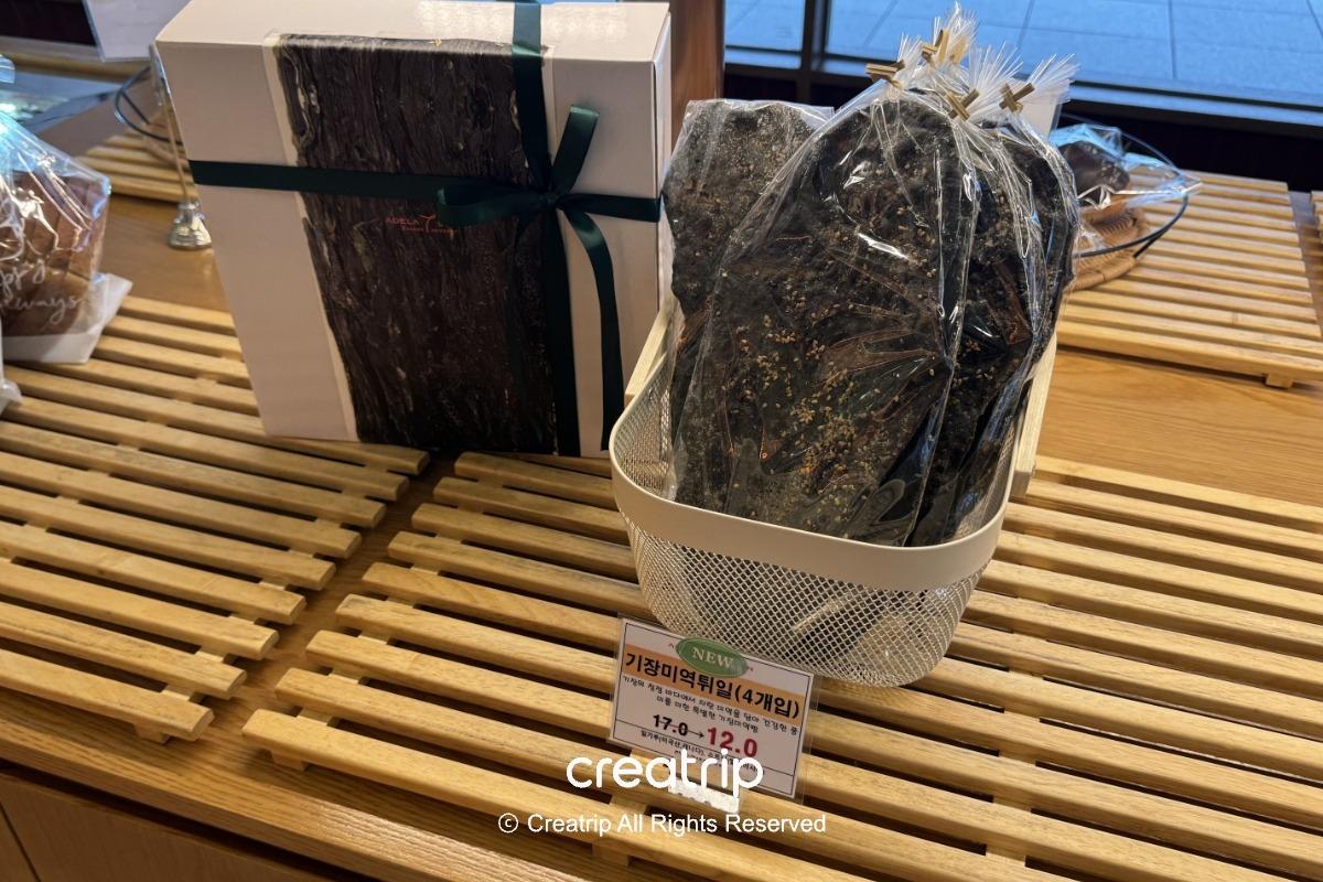ADELA 7的機張海帶脆餅包裝展示：透明袋裝海苔脆餅擺在白色籃子與木質陳列台上，旁邊有禮盒與價格標籤，適合作為金海機場伴手禮選擇