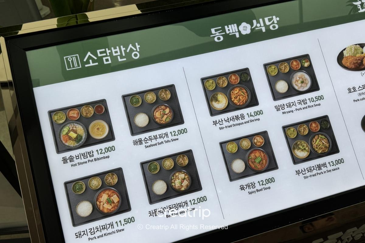 釜山金海机场餐厅电子菜单展示多款定食与价格，照片示范石锅拌饭、海鲜豆腐锅、泡菜猪肉汤等韩式套餐，方便旅客点餐参考（含英文品名与价位）。