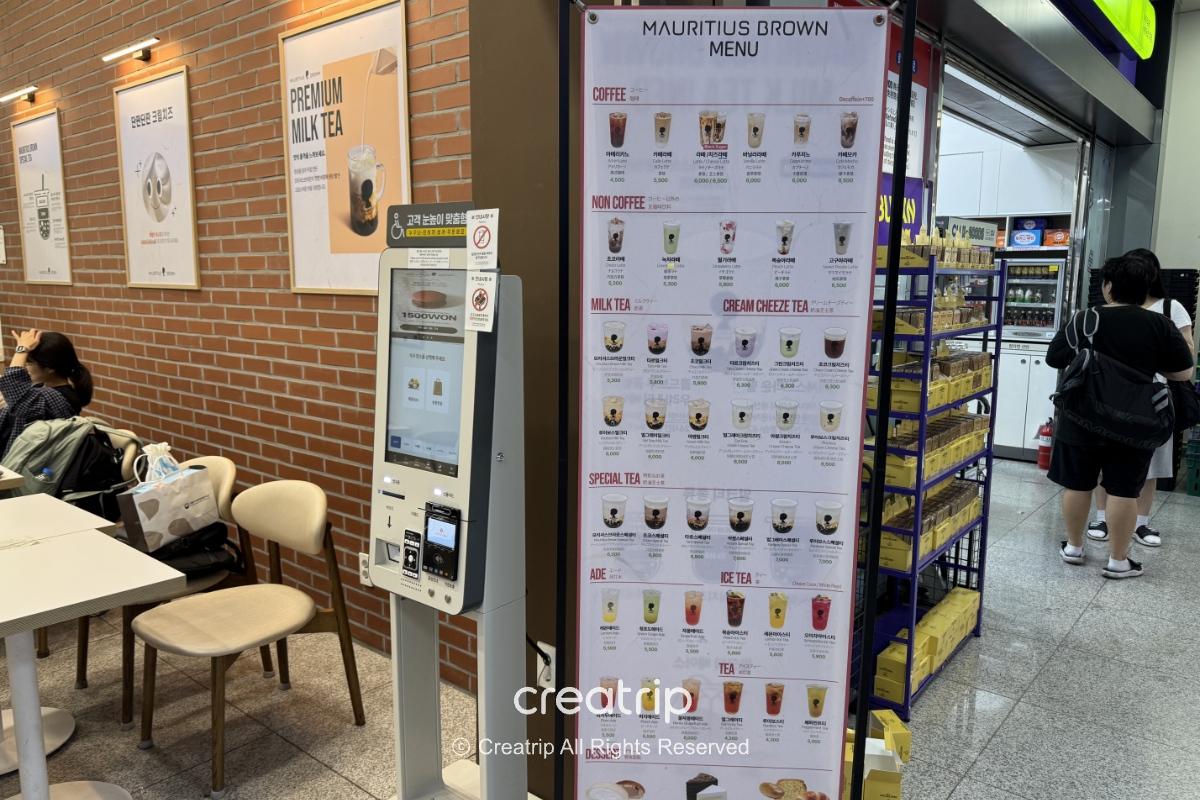 釜山金海机场国际线抵达层的Mauritius Brown柜位，展示大型菜单与自助点餐机，旁边有座位区和便利商店货架，适合下机买饮料或外带奶茶。