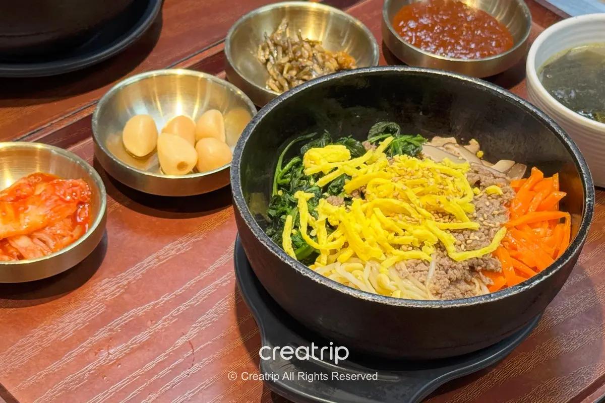 石鍋拌飯特寫：熱騰騰石鍋內有牛絞肉、炒菠菜、紅蘿蔔絲、豆芽與細切蛋絲，旁邊配有泡菜、小菜與醬料，呈現金海機場PLE美食街的韓式套餐風味